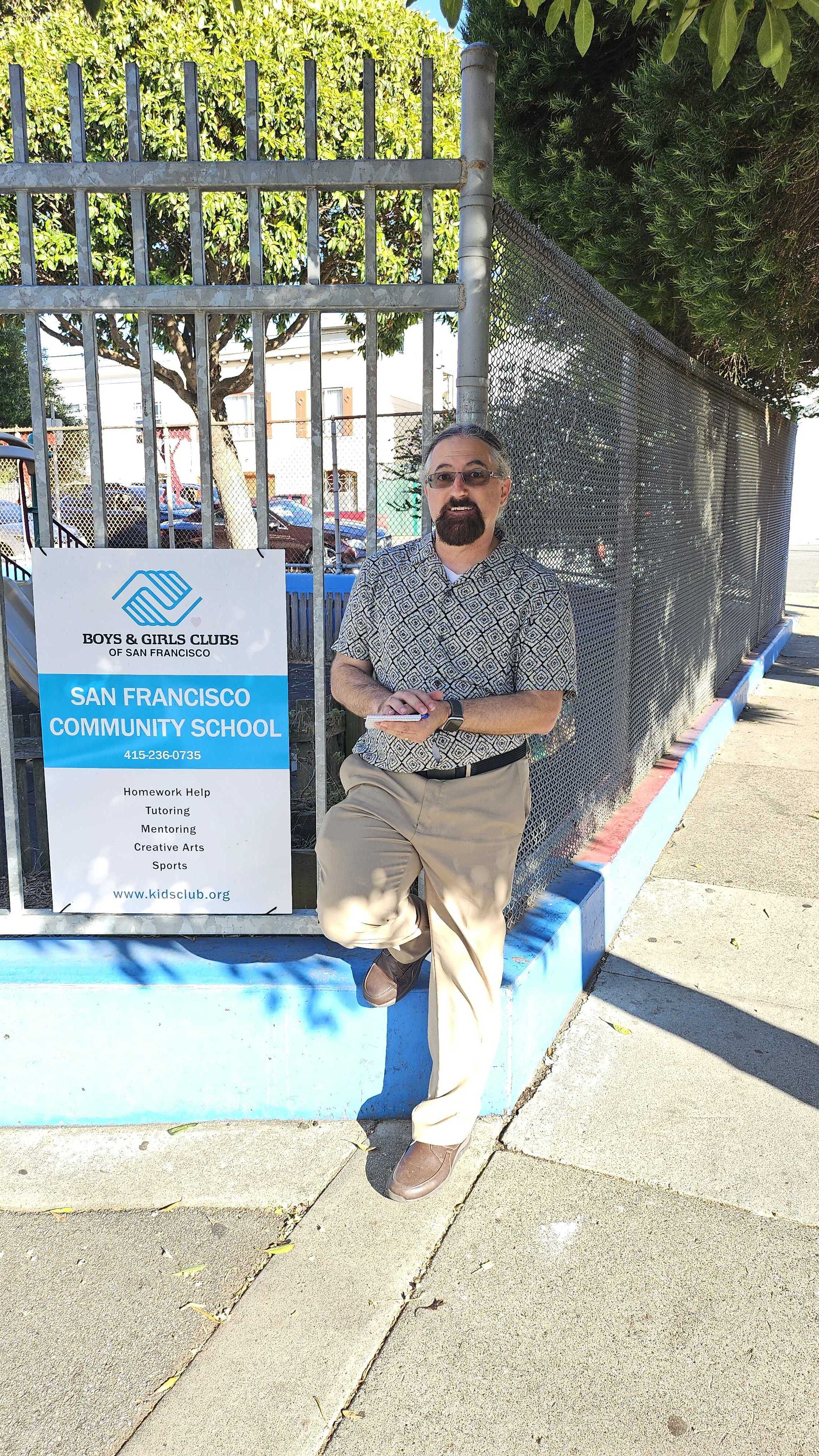 A man in a patterned shirt and khaki pants leans against a fence beside a sign for the San Francisco Community School's Boys &amp; Girls Clubs, with trees around.