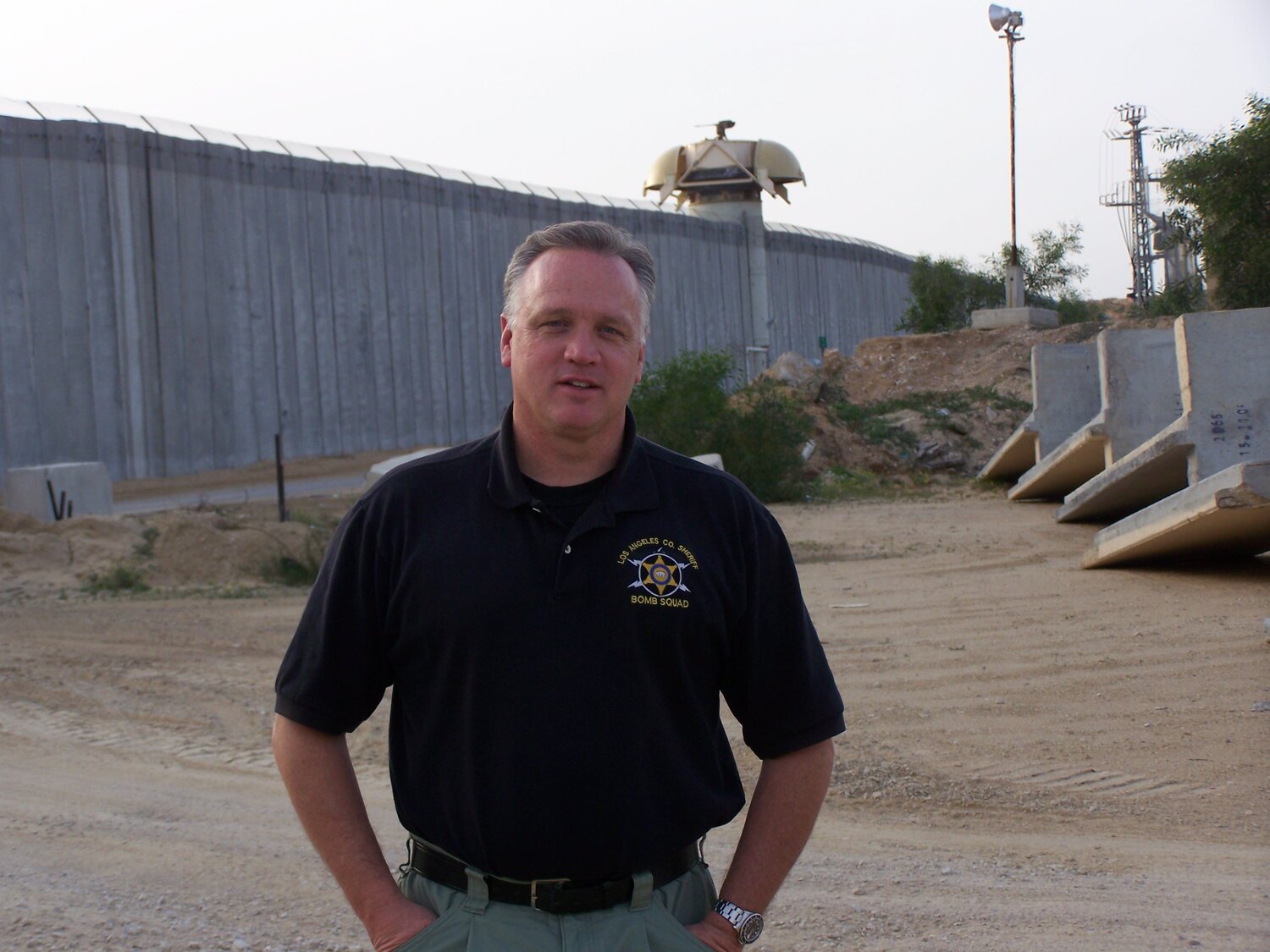 A man in a black shirt stands on a dirt path in front of a tall concrete wall. There are some barriers and sparse vegetation around him.