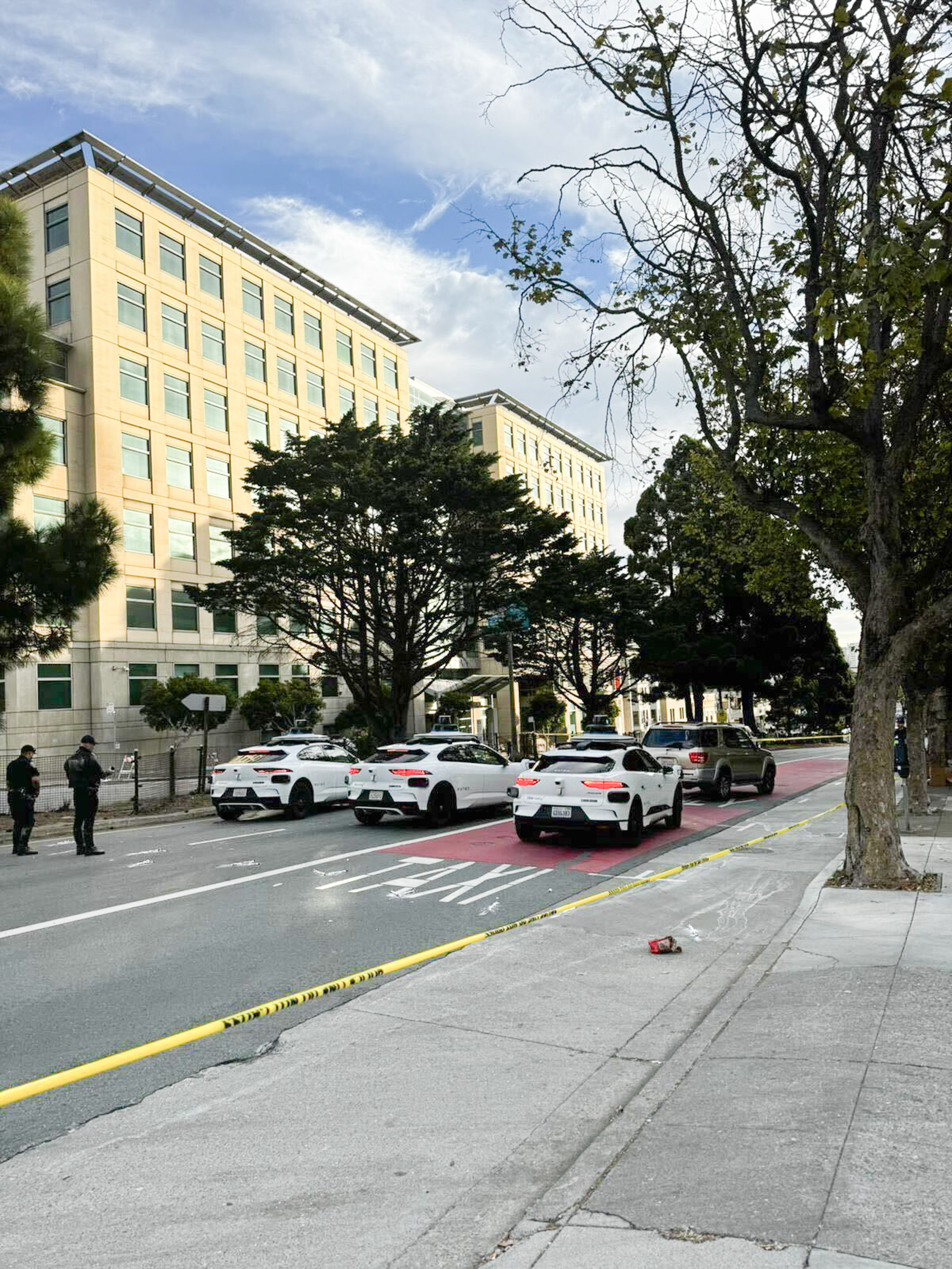 A street is cordoned off with yellow tape, lined with police cars and officers. Tall buildings and trees are in the background under a partly cloudy sky.