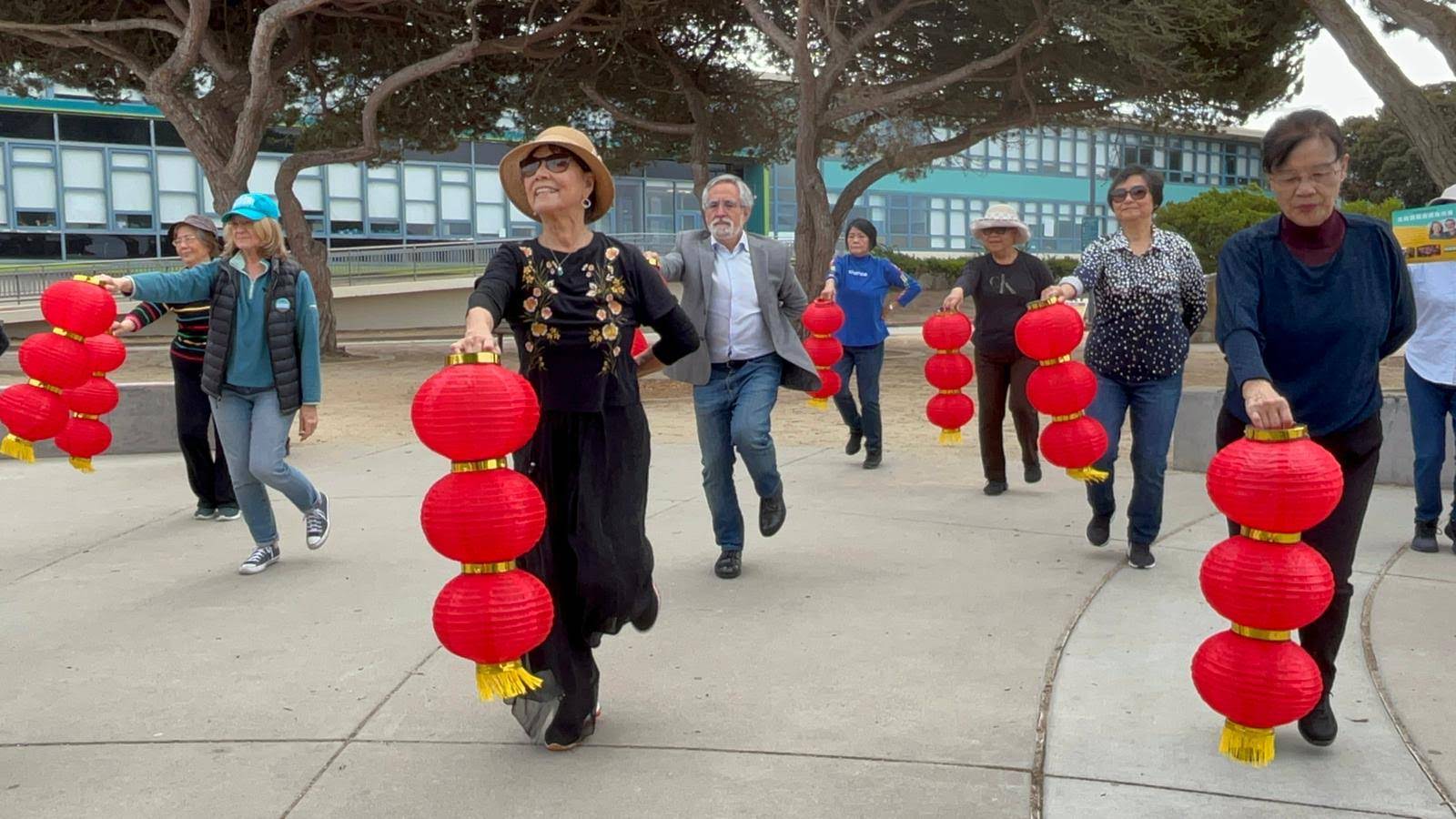 A group of people joyfully walk while holding red lanterns outdoors. The backdrop includes trees and a building. Everyone appears engaged and cheerful.