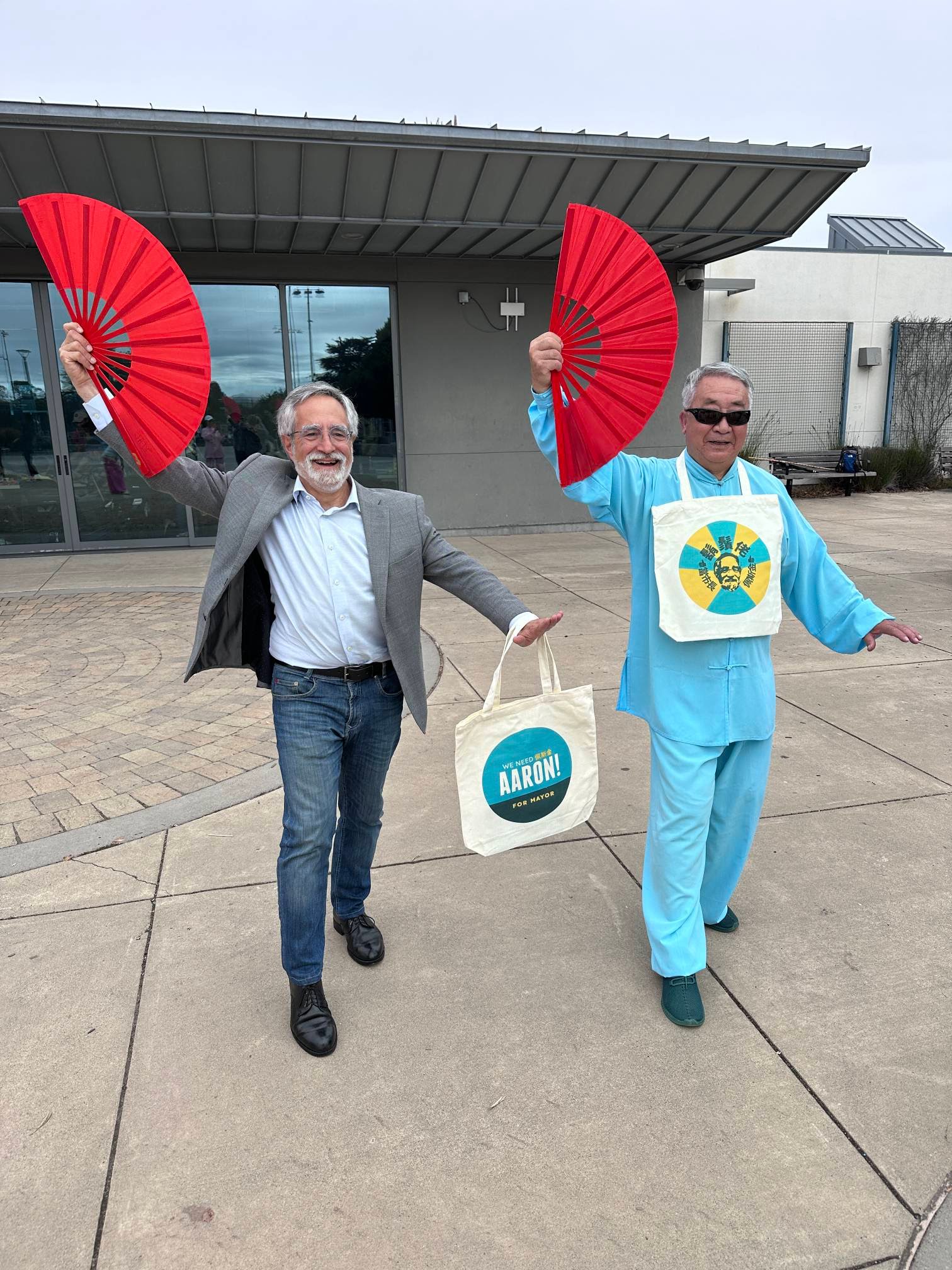 Two men smile and hold red fans. One wears a suit with jeans and carries a tote bag saying &quot;AARON! for Mayor.&quot; The other wears bright blue traditional attire.