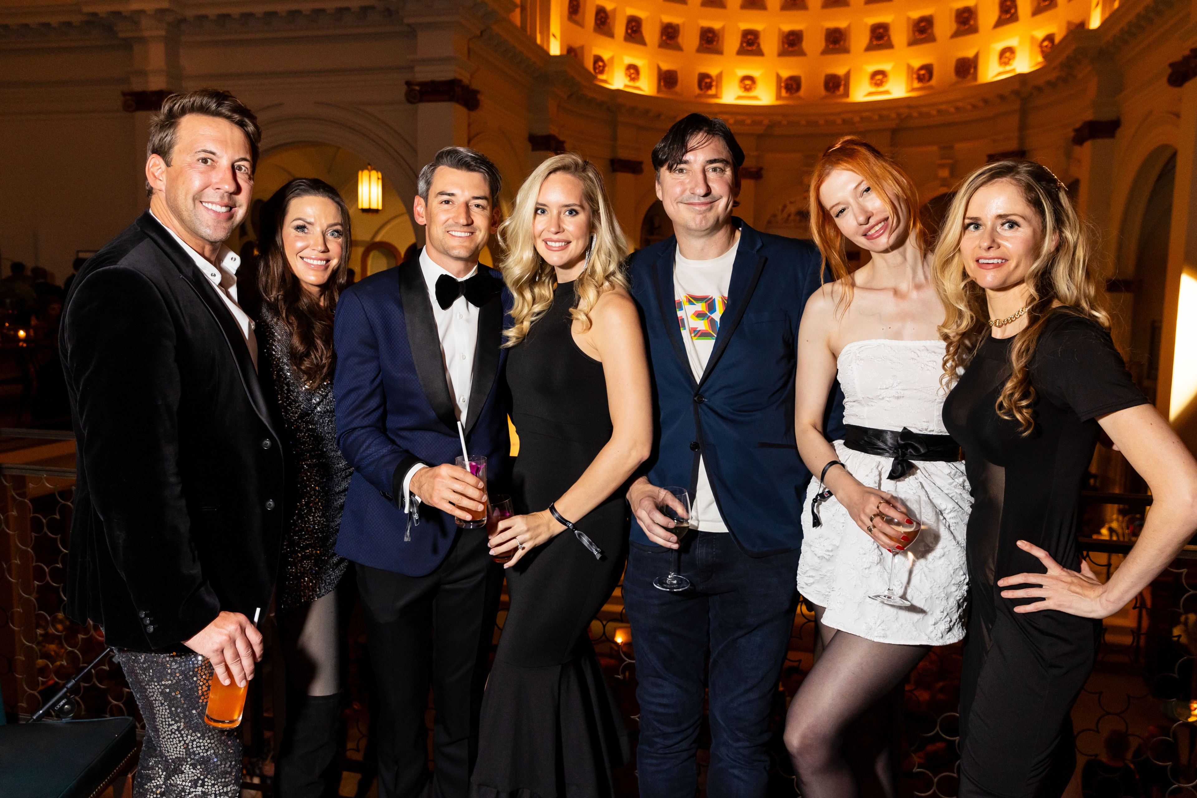 A group of seven well-dressed people are posing together at an upscale event, holding drinks. The background is dimly lit, featuring elegant architecture.