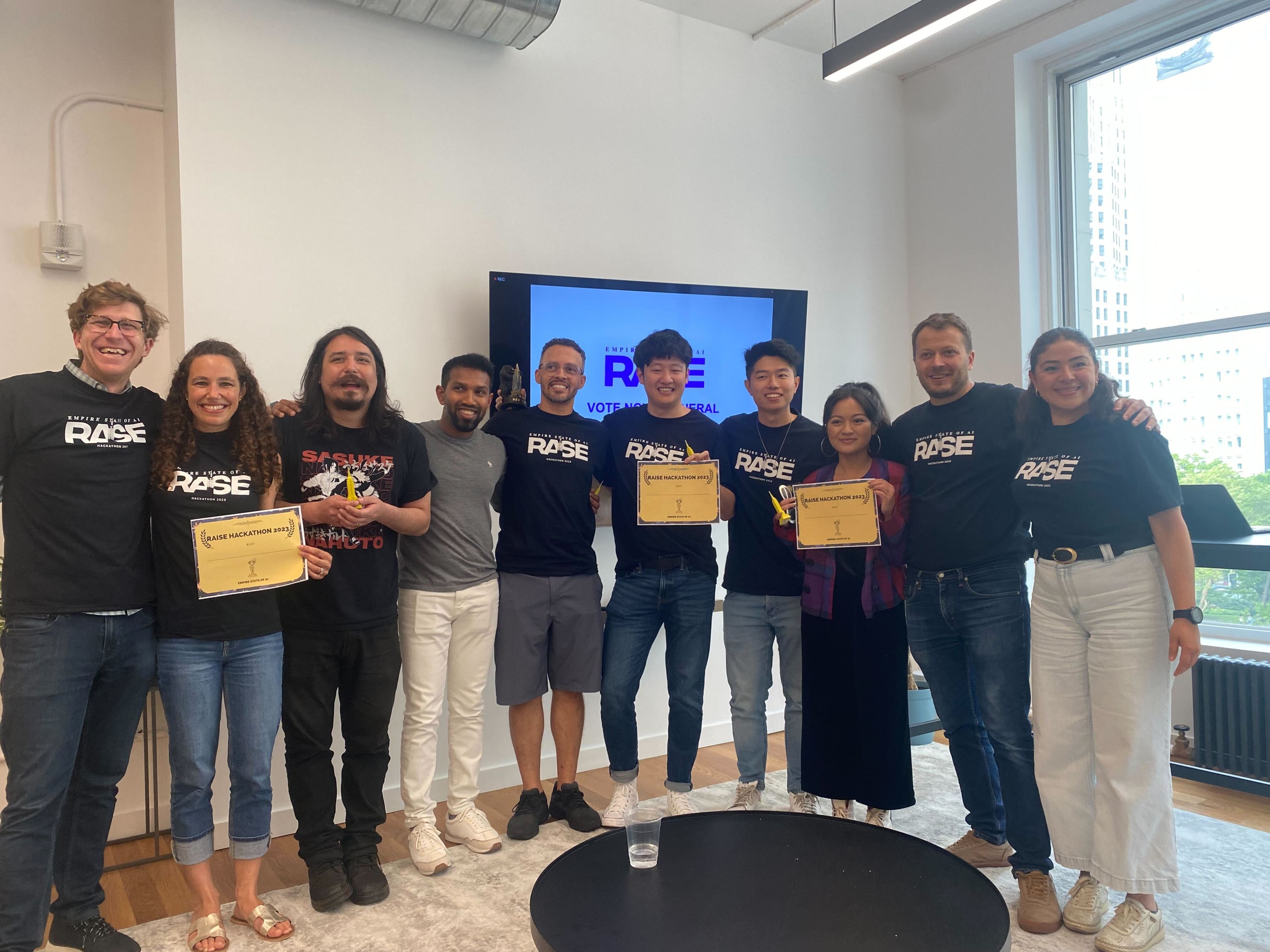 A group of smiling people stands indoors, holding certificates and a trophy. Most wear matching black shirts, and a screen in the background displays &quot;RAISE HACKATHON 2023.&quot;