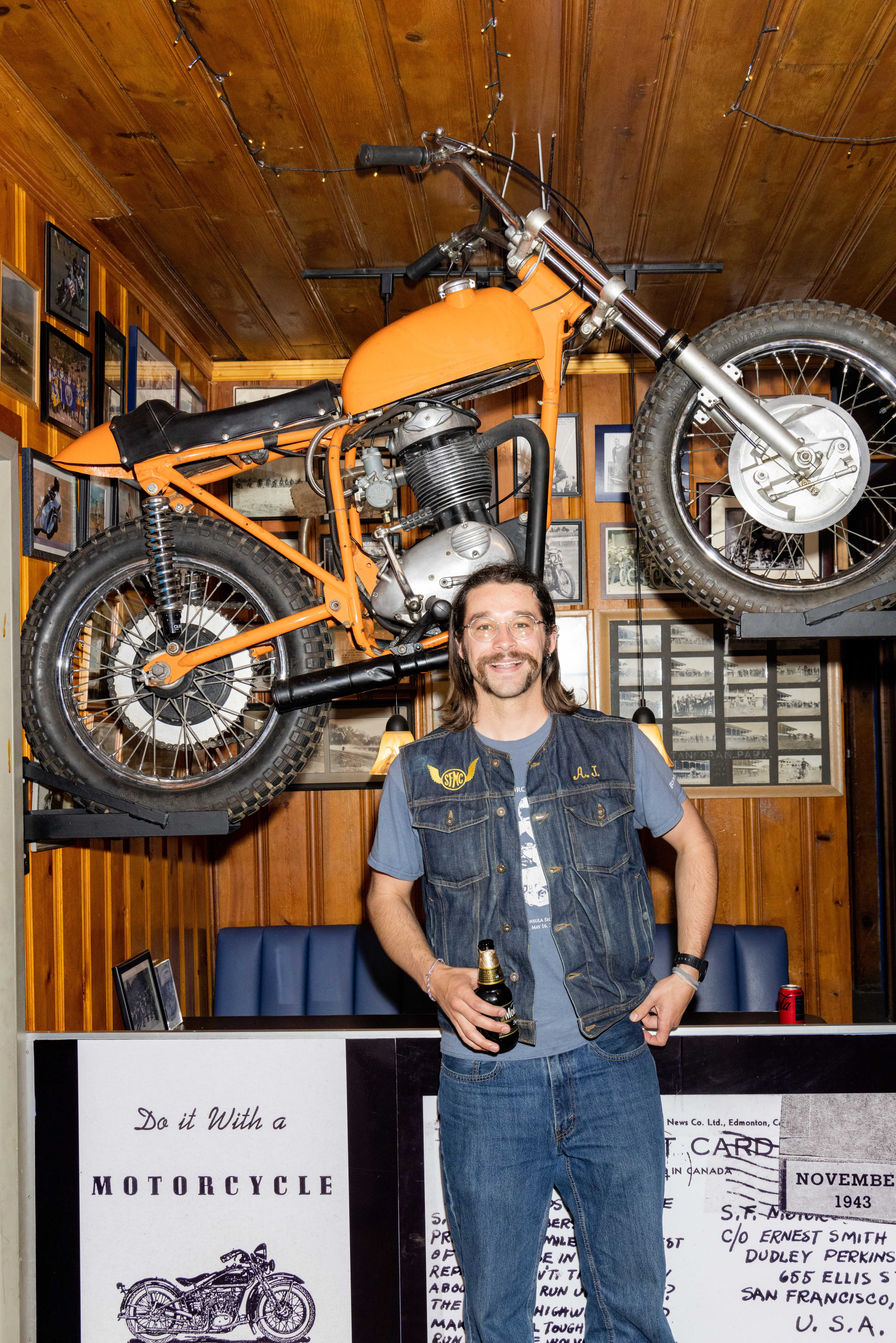 A smiling man with long hair stands holding a bottle, wearing a denim vest and blue jeans. Behind him, an orange motorcycle is mounted on a wall with pictures.