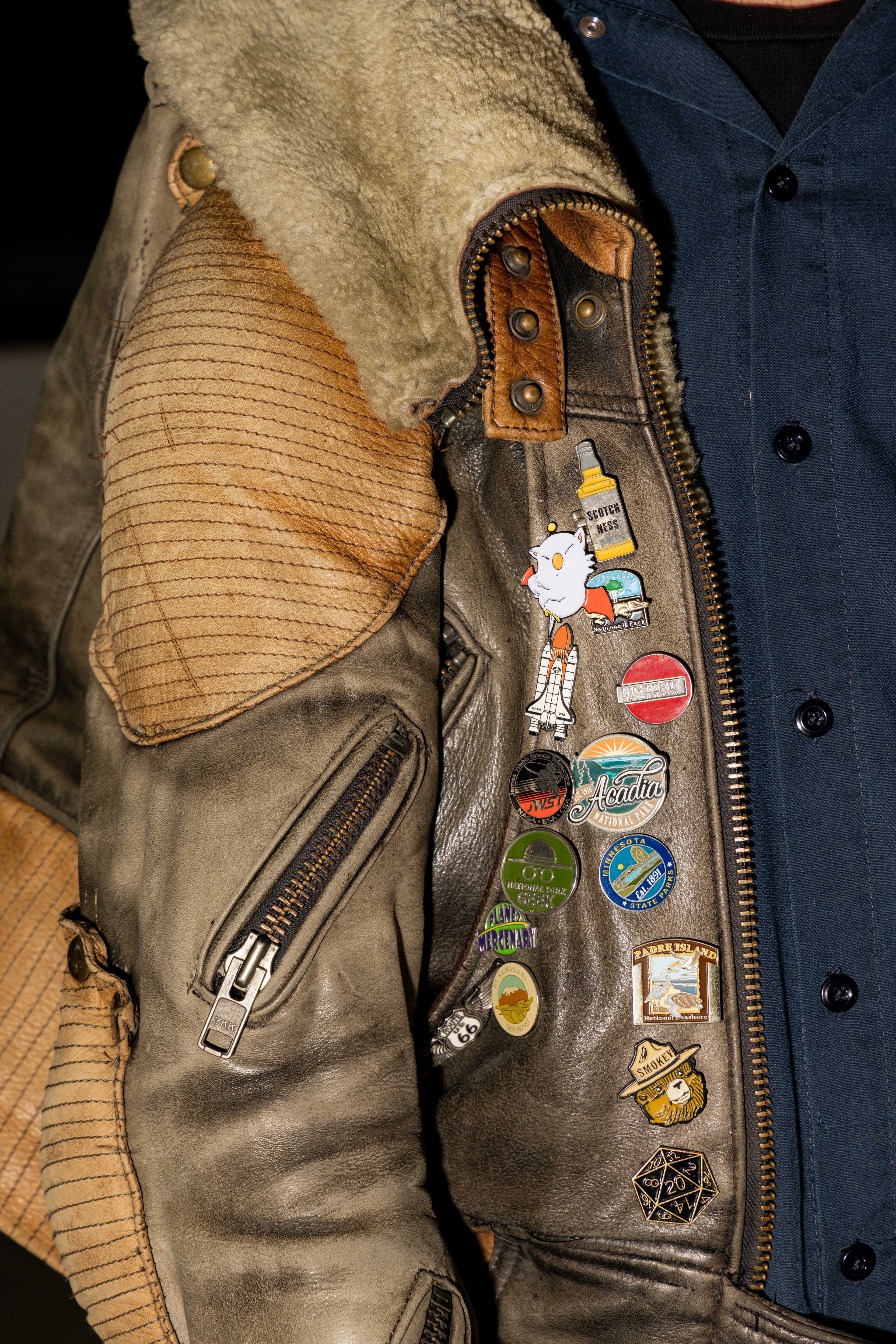 A worn leather jacket with a fur collar displays various colorful pins, including a bottle labeled &quot;Scotch Ness&quot; and other travel-themed designs.