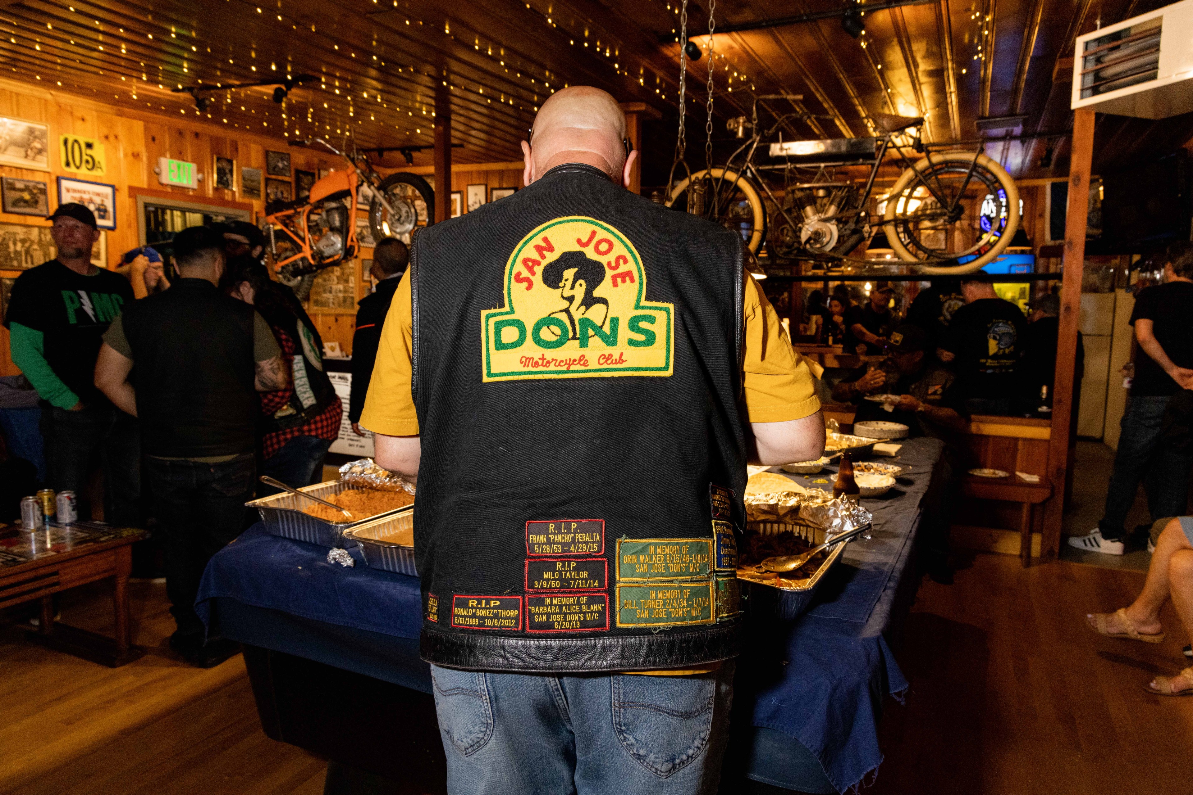 A person wearing a &quot;San Jose Dons Motorcycle Club&quot; vest stands by a food table at a dimly lit gathering, with vintage motorcycles displayed on the walls.