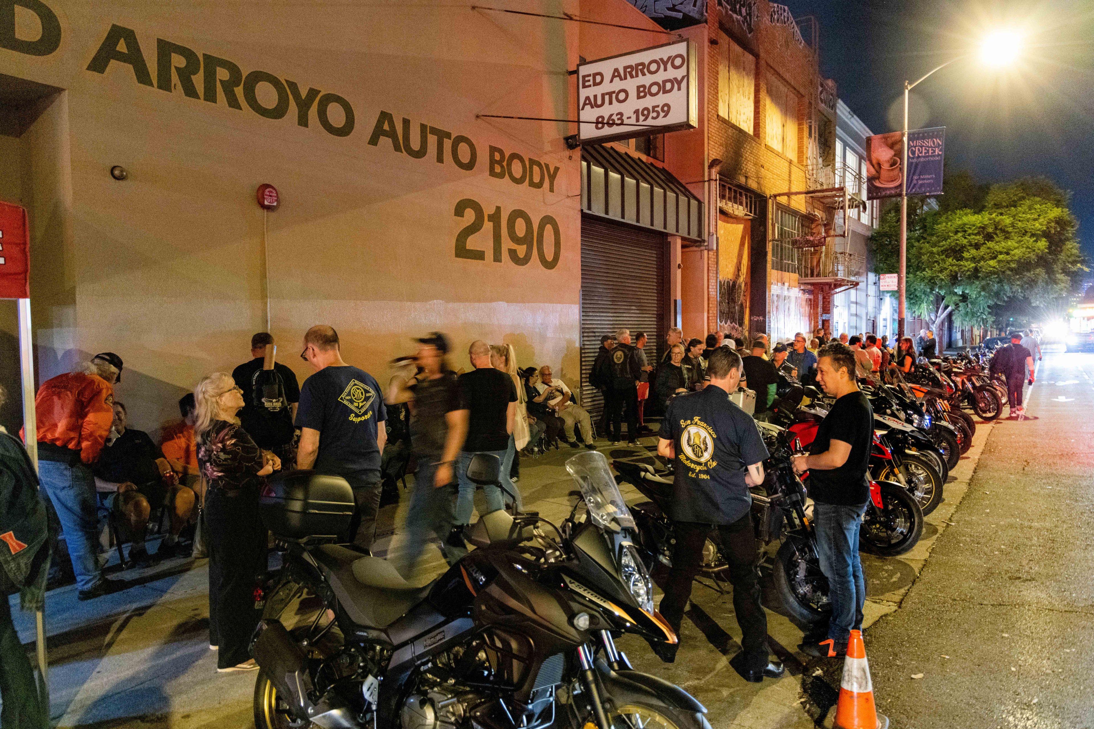 A group of people gathers outside &quot;Ed Arroyo Auto Body,&quot; with numerous motorcycles lined up along the street. The scene is illuminated by warm streetlights.