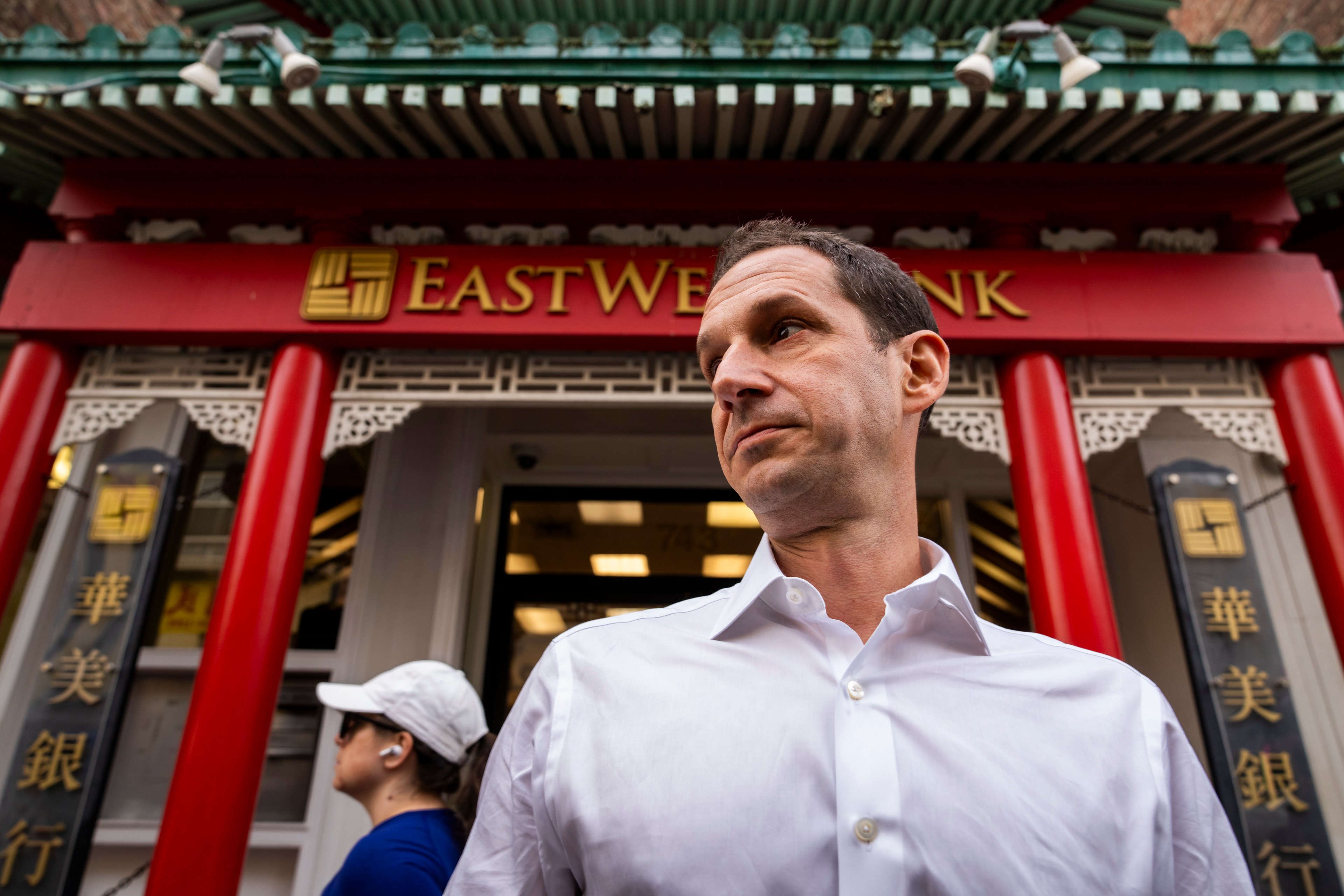 A man in a white shirt stands outside East West Bank, featuring traditional architectural elements with red pillars and a green roof. Another person is in the background.