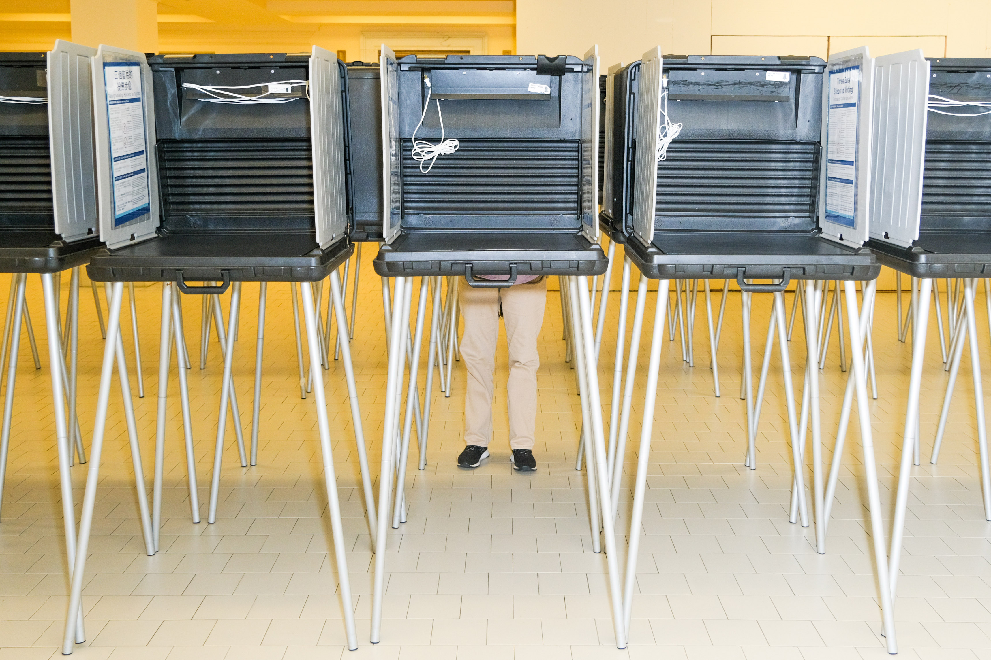 A row of empty voting booths is set up in a brightly lit room. Only a person's legs are visible beneath one booth, indicating someone using it.