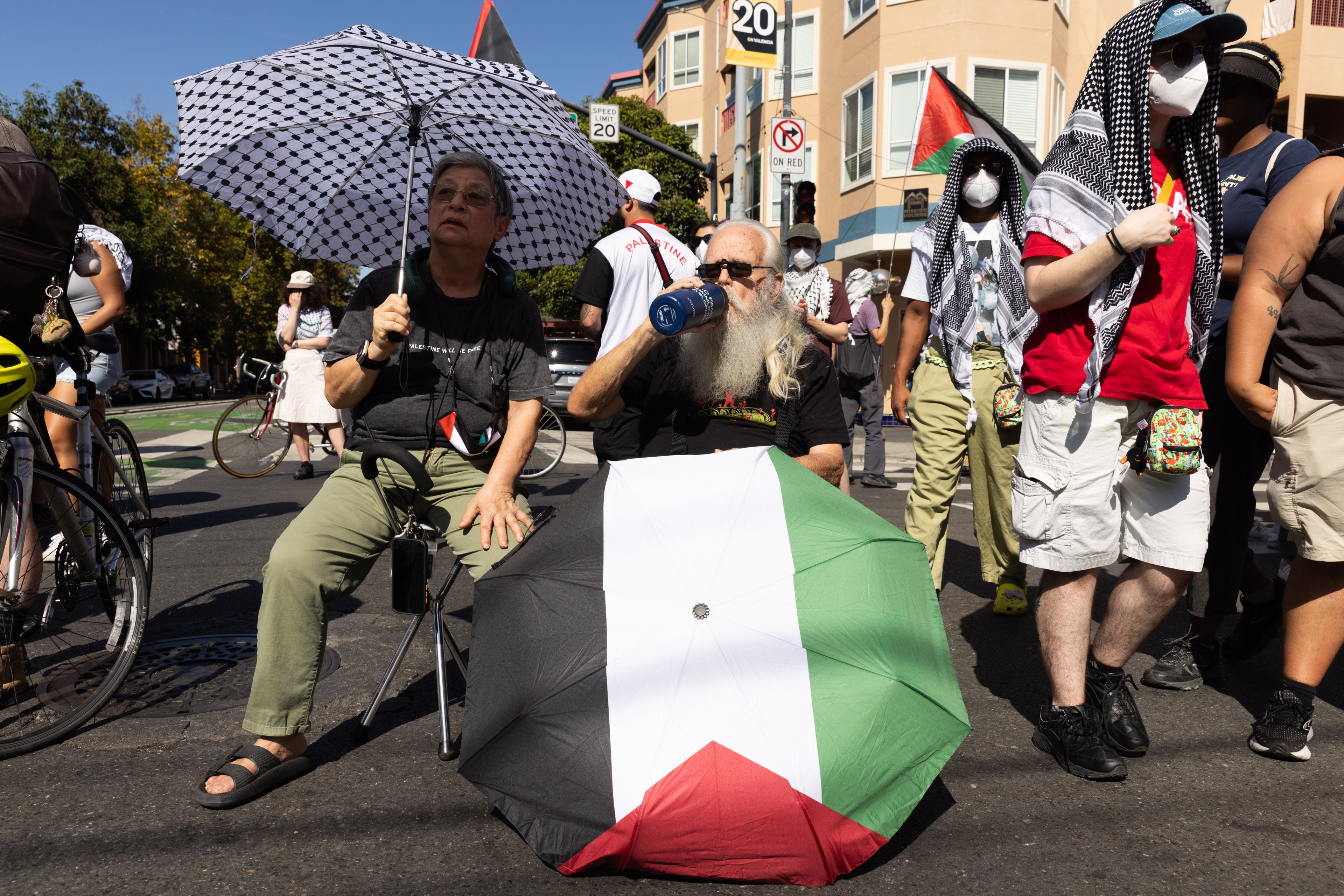 People sit and stand on a street, one holding a checkered umbrella, another with a Palestine flag umbrella. Others wear keffiyehs and masks, suggesting a protest.