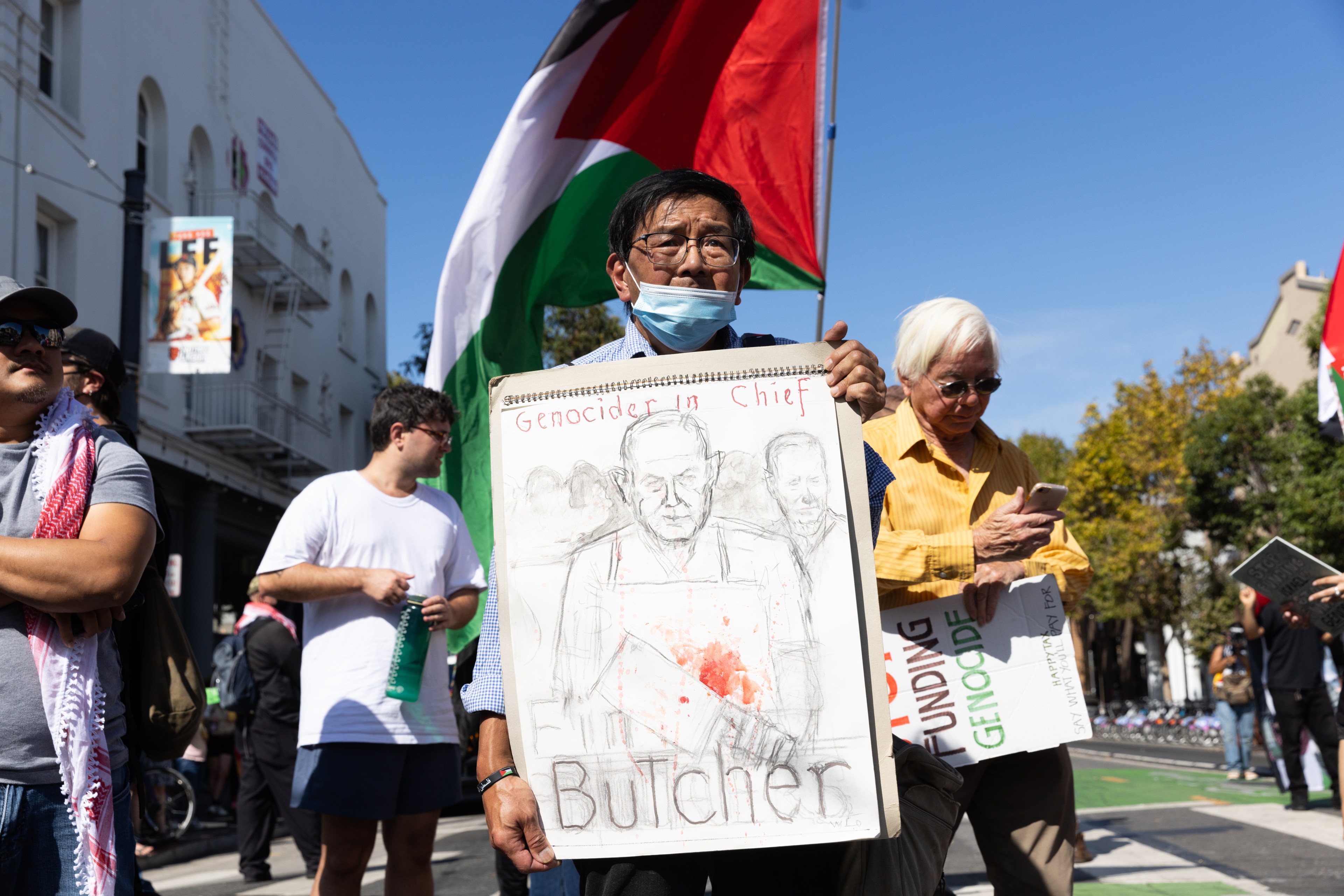 A man holds a sign with a sketch labeled &quot;Genocider in Chief&quot; and &quot;Butcher&quot; at a protest. Others nearby hold signs, and a large Palestinian flag is in the background.