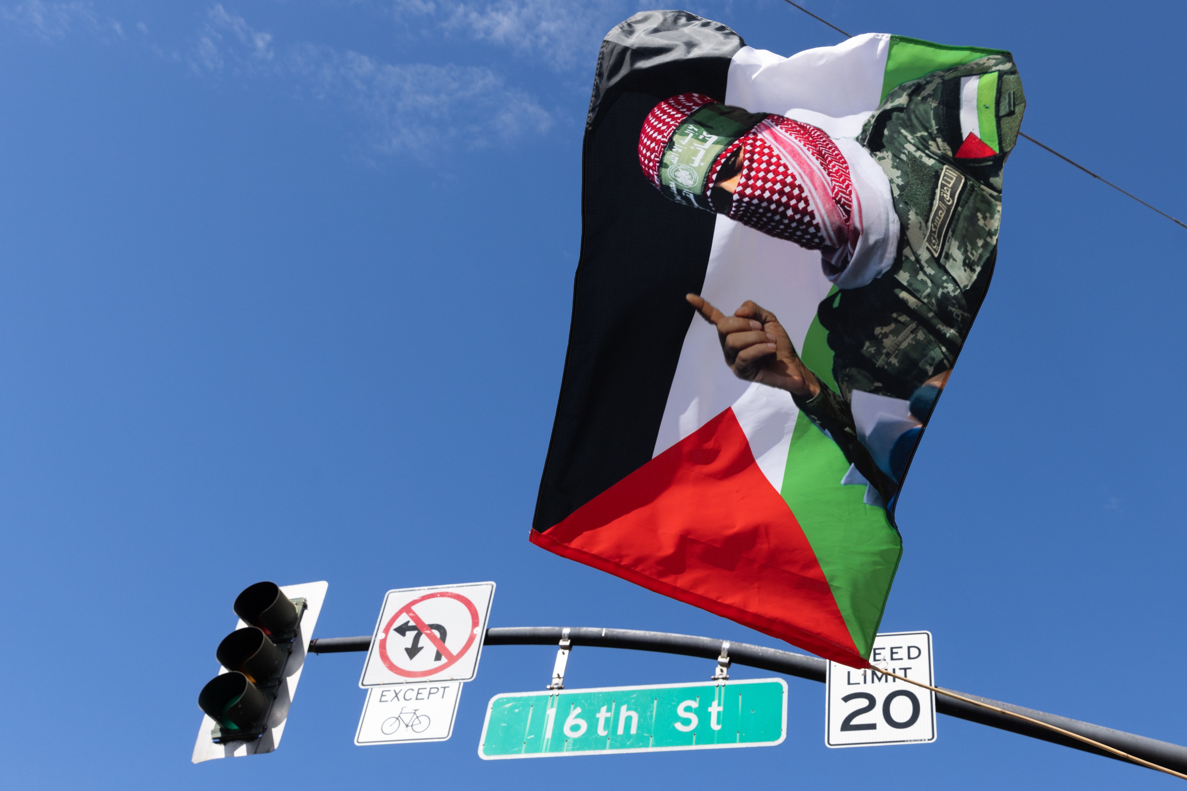 A flag featuring a masked figure is attached to a streetlight, with clear skies in the background. Below are traffic signs and a 16th St street sign.