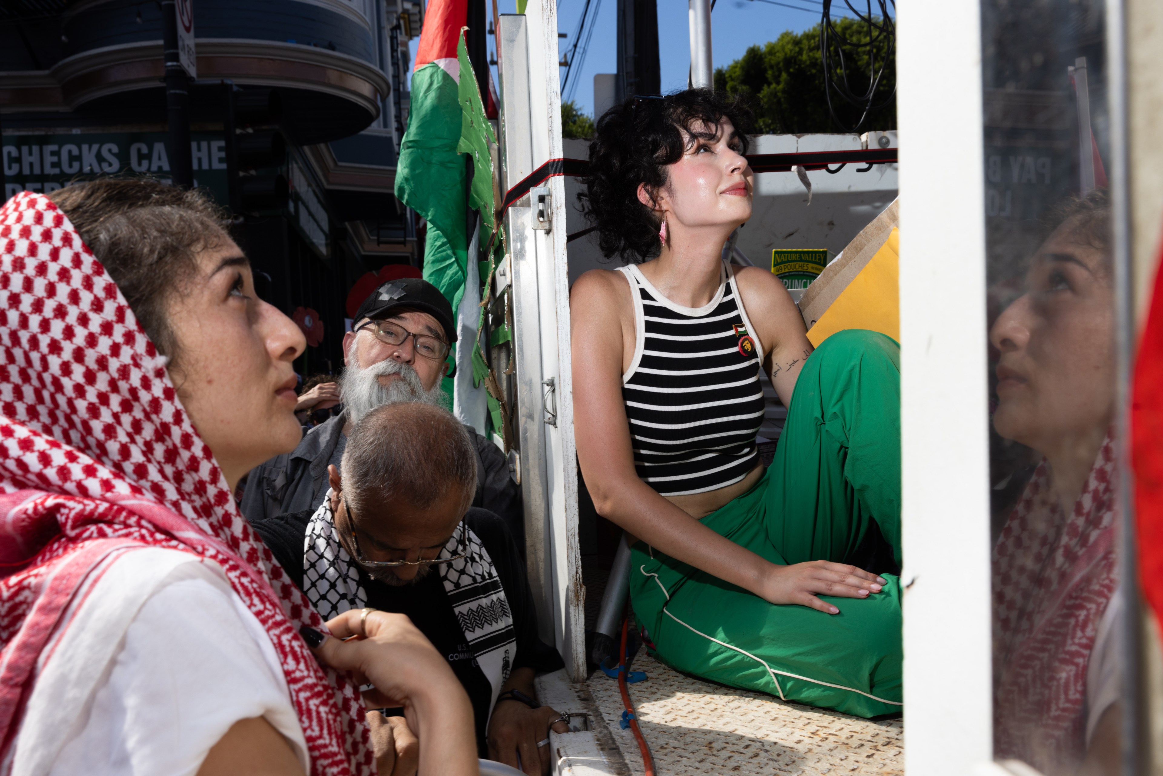 A group of people gather closely in an urban setting, displaying a colorful flag. They appear focused and engaged, with diverse clothing styles and expressions.