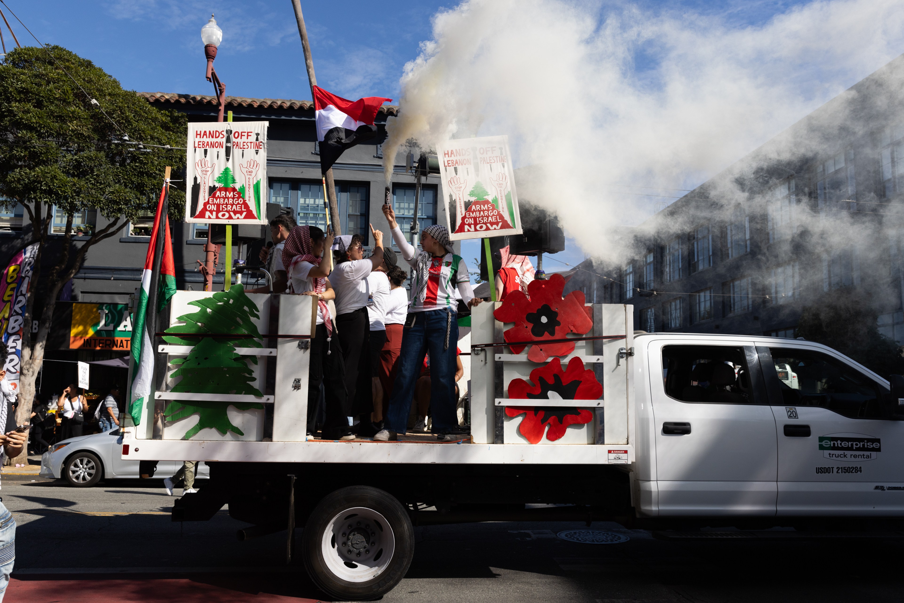 A white truck carries people holding protest signs and waving flags, with smoke in the air. Decorations include green and red symbols, and “Hands Off Lebanon &amp; Palestine” is visible.