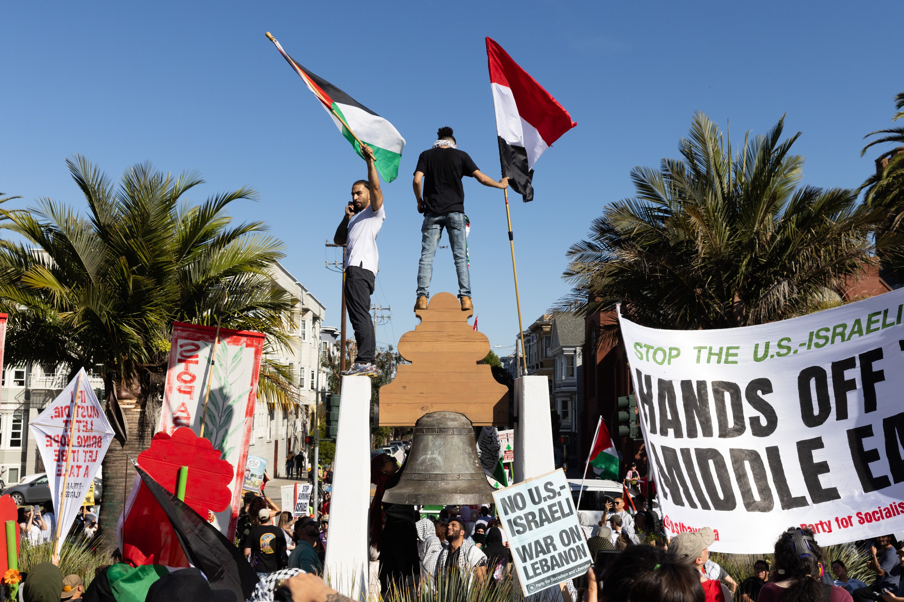 A group of protesters holds flags and signs with messages against U.S.-Israeli actions. Two men stand elevated, waving flags, among palm trees and banners.