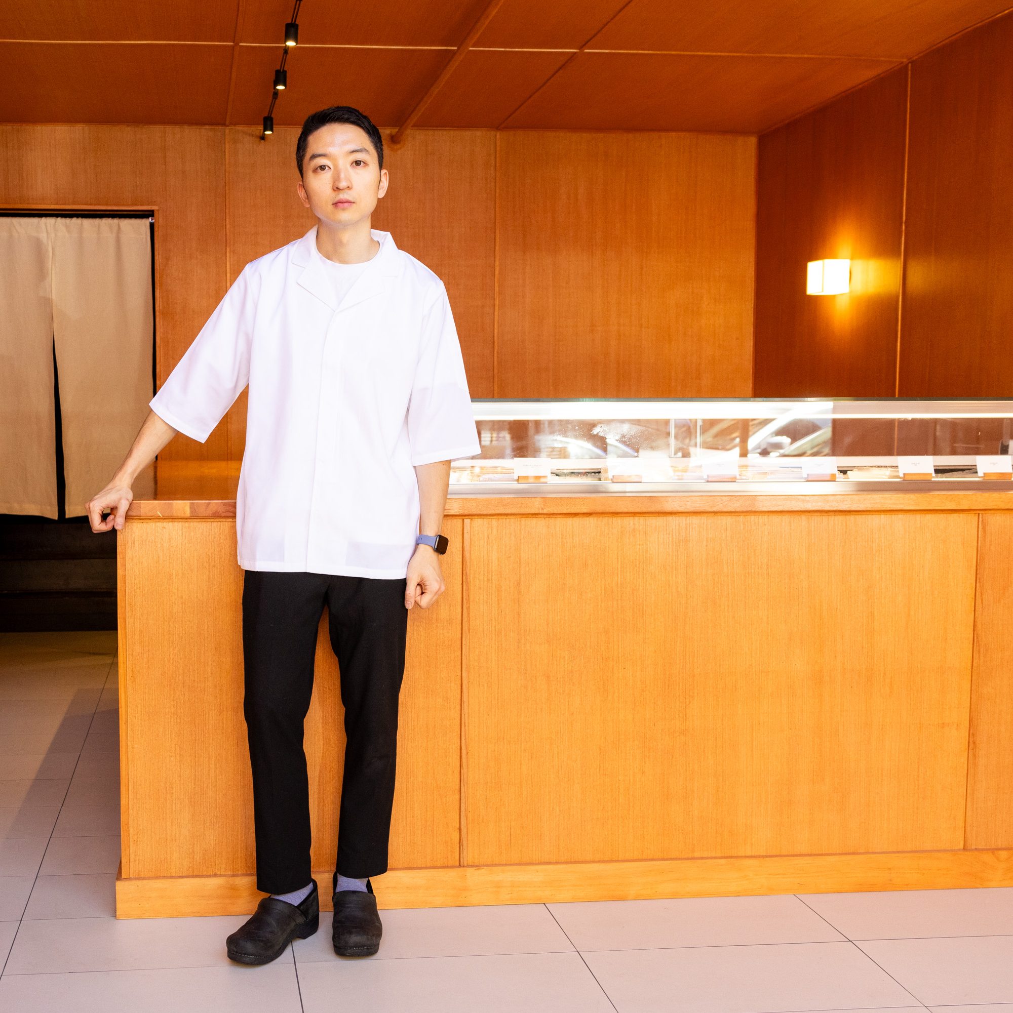 A person in a white shirt and black pants stands beside a light wooden counter. The room has wooden walls and a small light fixture.