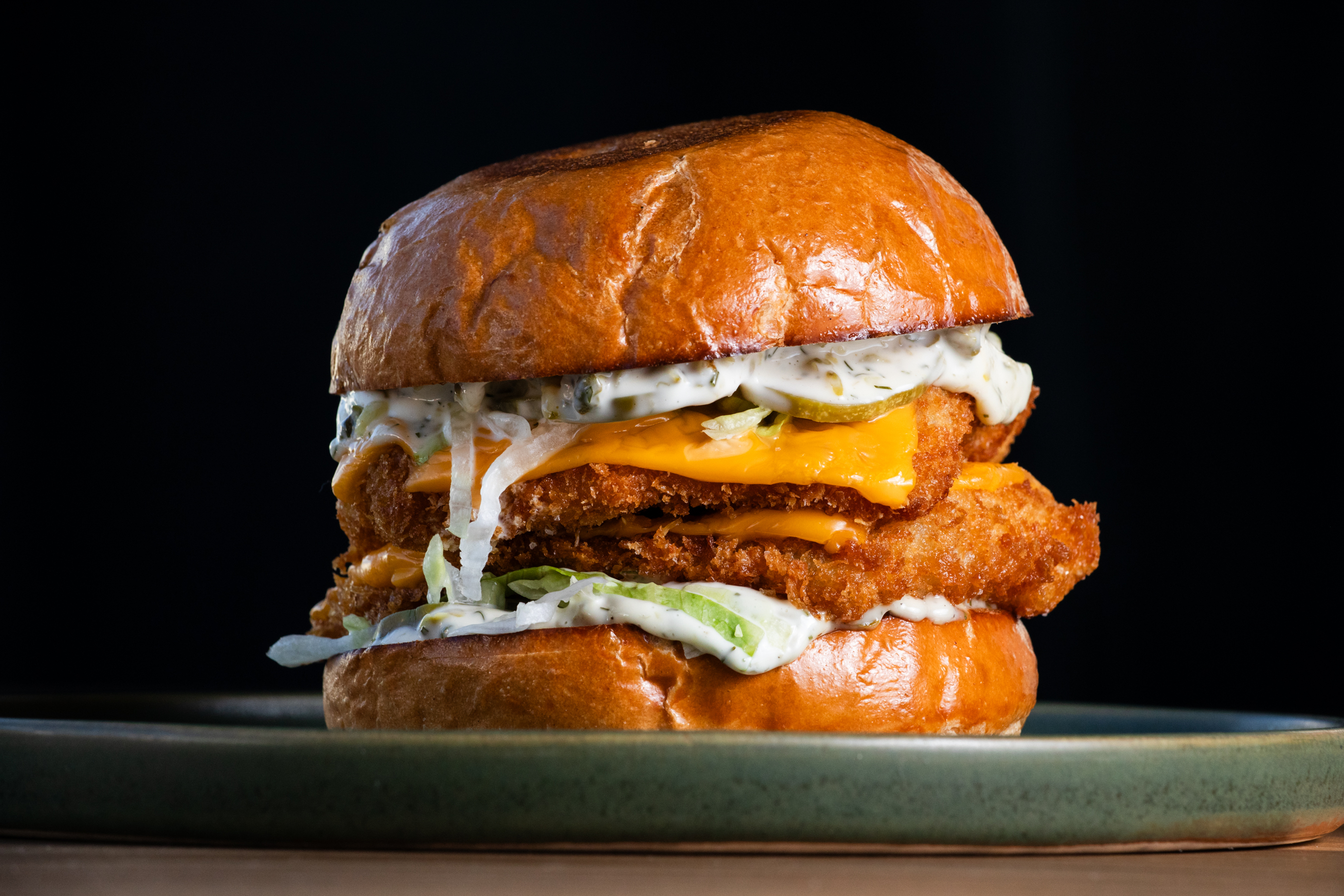 A crispy chicken sandwich with melted cheese, pickles, and creamy sauce is layered between a shiny, toasted bun on a plate against a dark background.