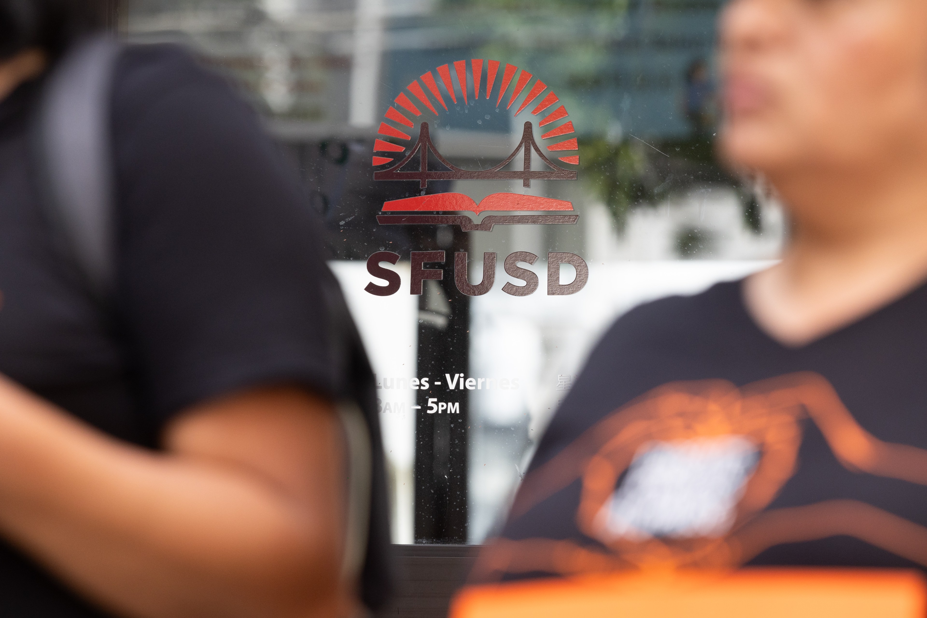 Two people are partially visible in front of a glass door with an SFUSD logo, featuring a bridge, book, and sun design.