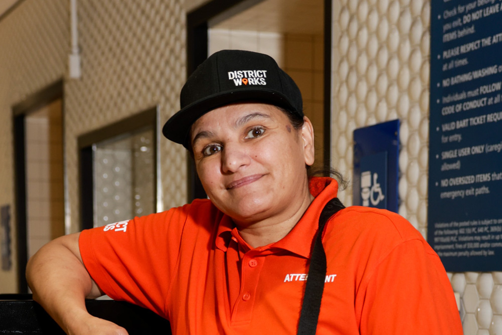 A person in a red shirt and black cap labeled "District Works" sits smiling, holding a device. They appear to be an attendant in a well-lit setting.