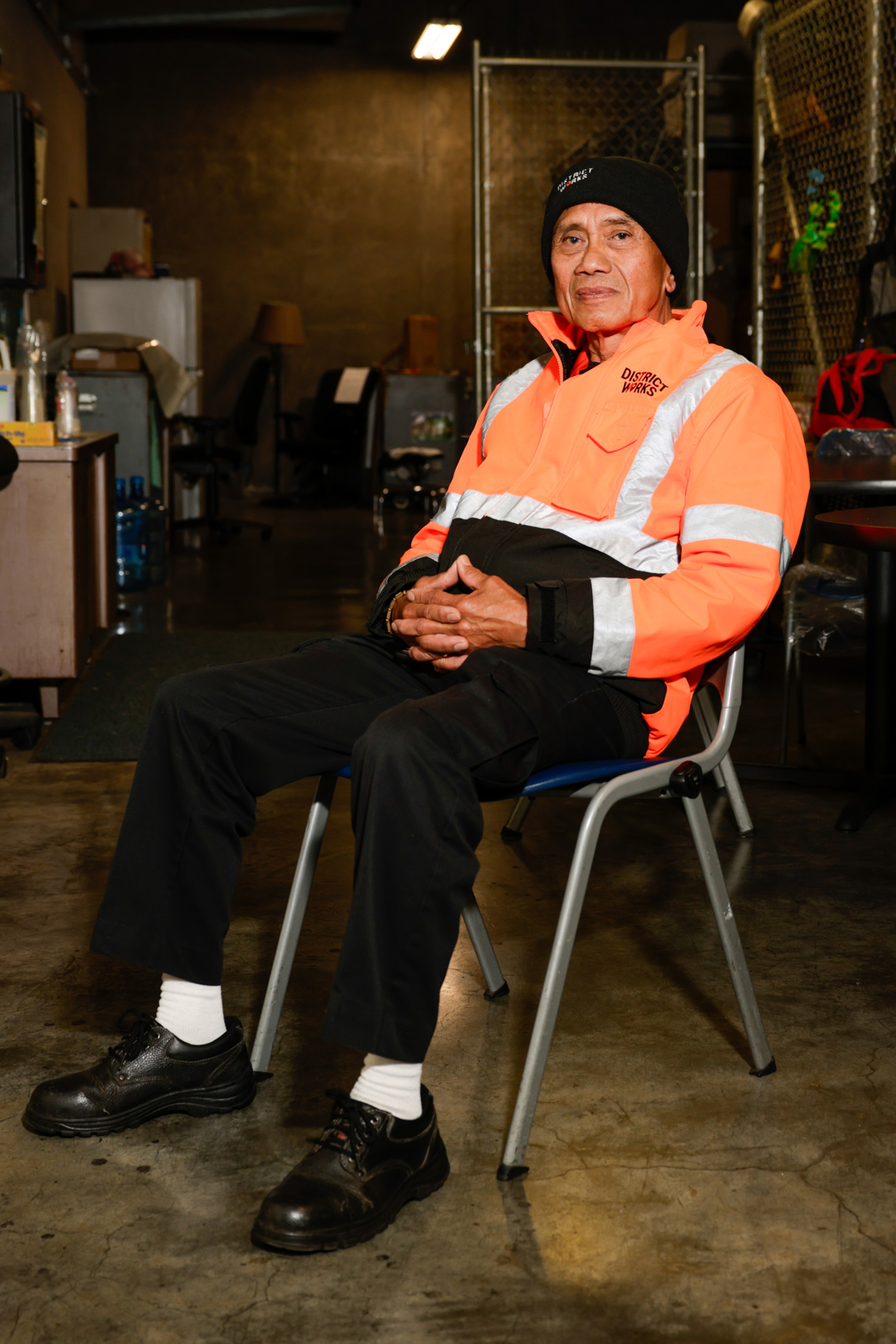 A person in a bright orange safety jacket and black beanie sits on a chair in a dimly-lit room, with hands clasped and a calm expression.