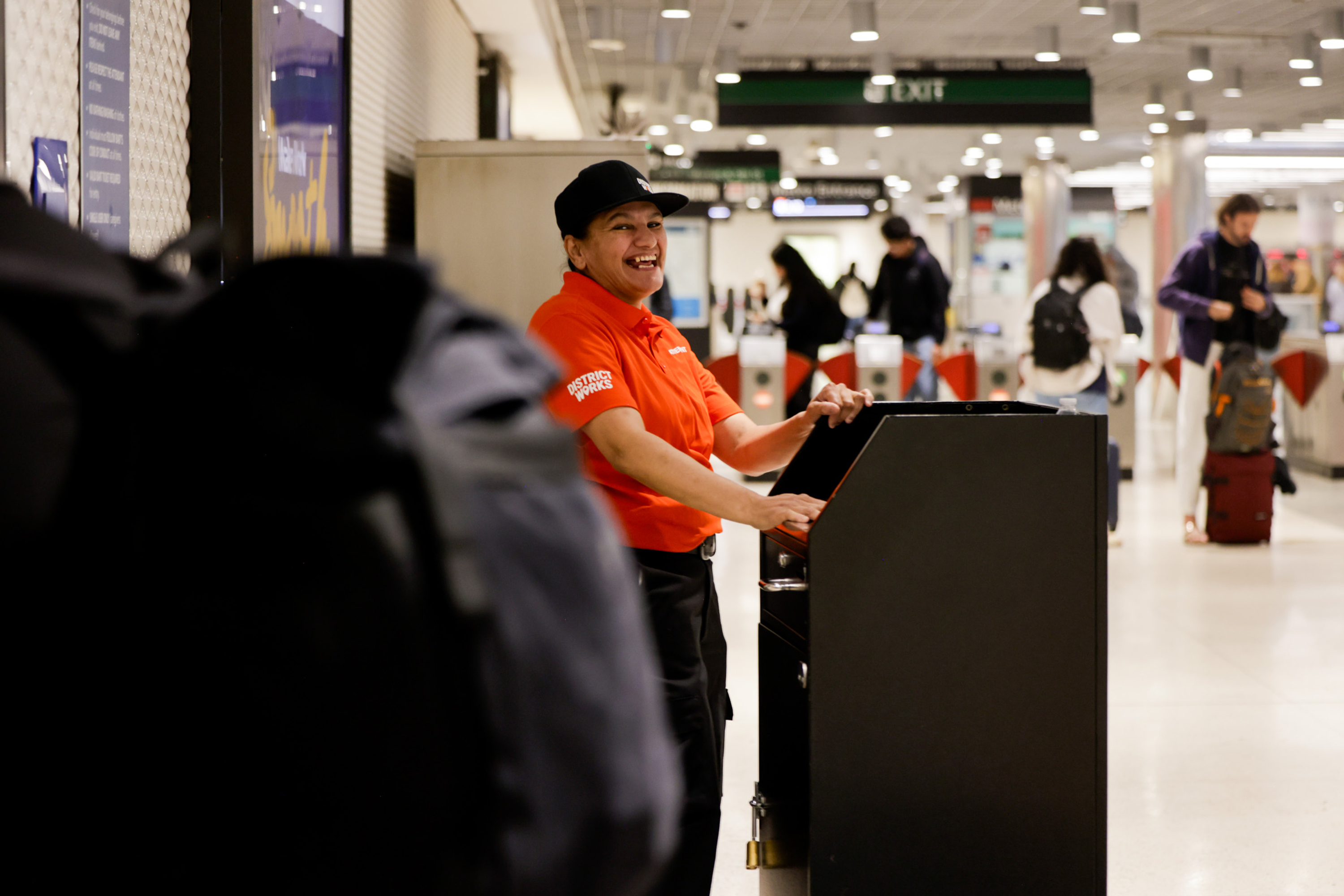 A person in a red polo shirt and cap smiles while handling a control unit in a busy subway station. People with luggage are in the background.