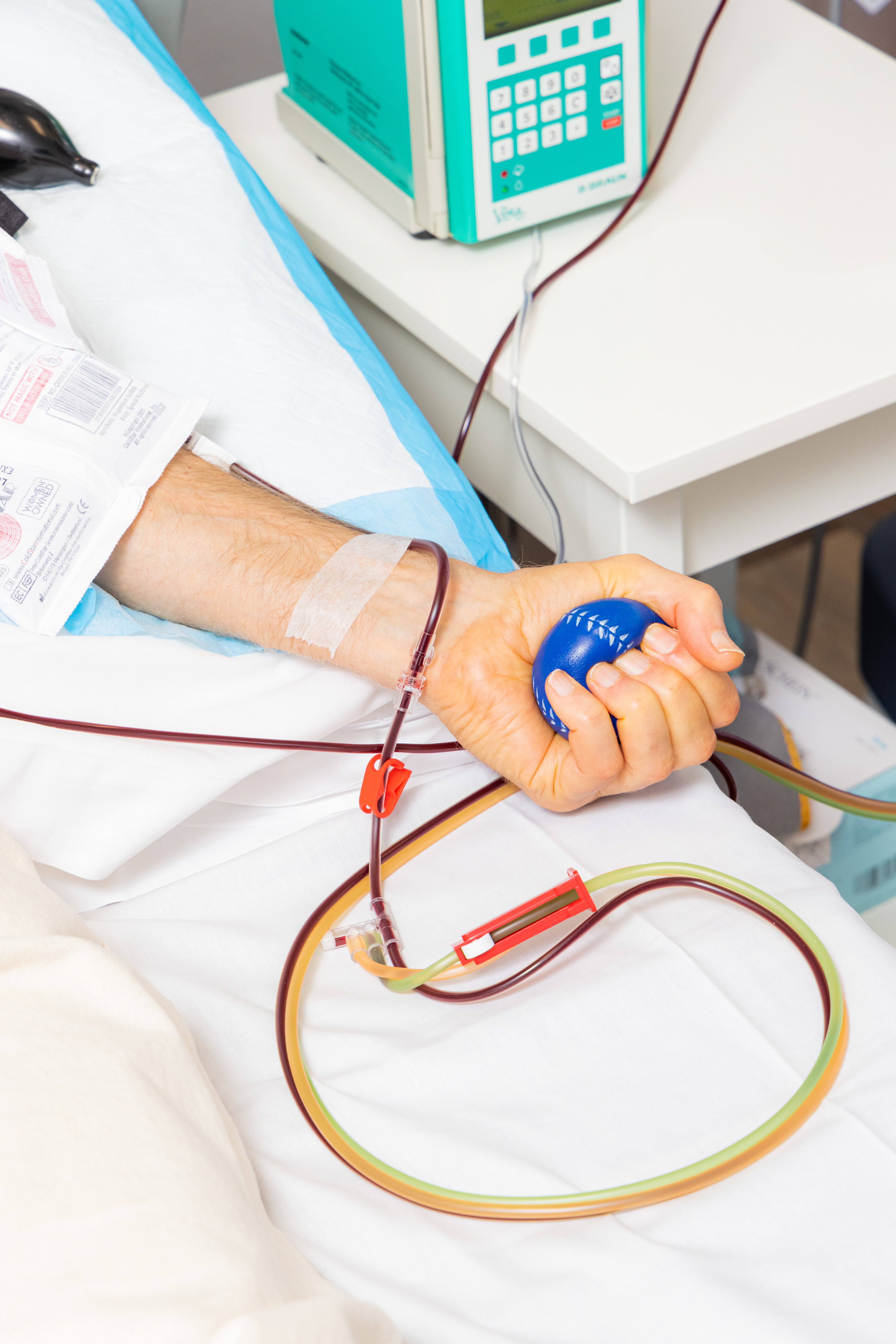 A person donates blood, seen by a tube attached to their arm, while squeezing a blue stress ball. A medical device is placed nearby.