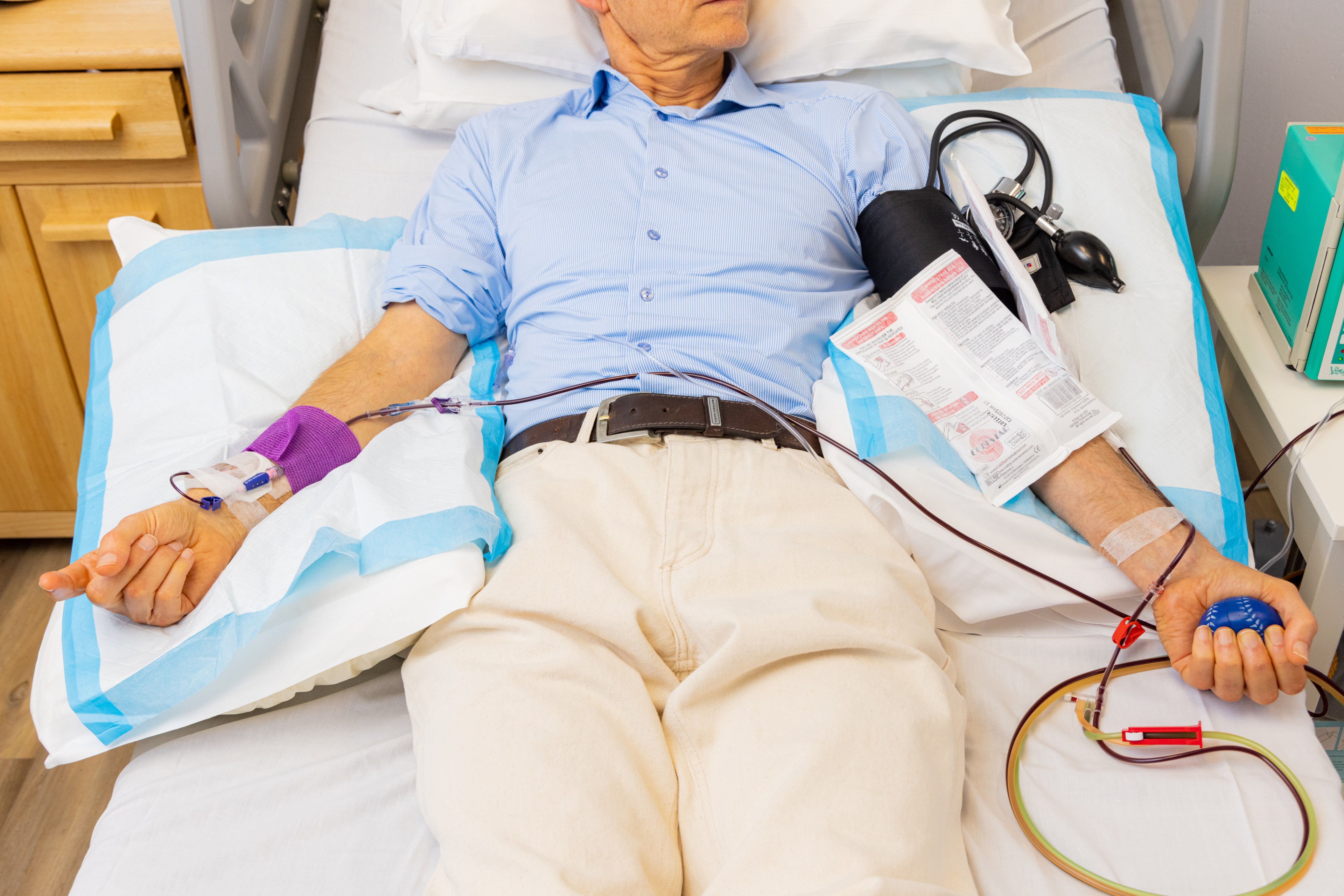 A person lies on a hospital bed donating blood, with a tube connected to their arm. They hold a blue stress ball and have a blood pressure cuff on one arm.