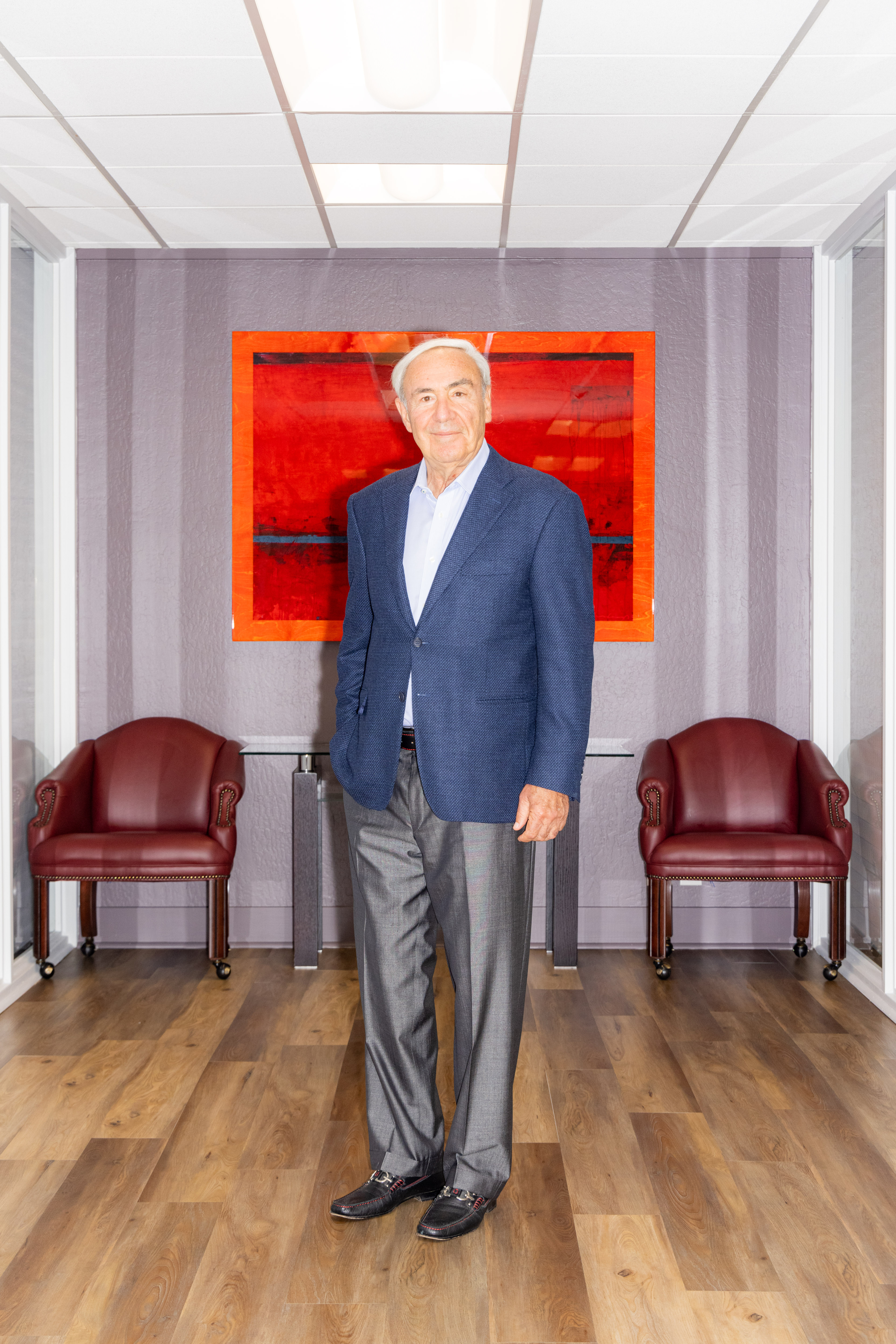 A man in a blue blazer and gray trousers stands in an office with wooden floors, flanked by two red chairs. A large red abstract painting hangs on the grey wall behind him.