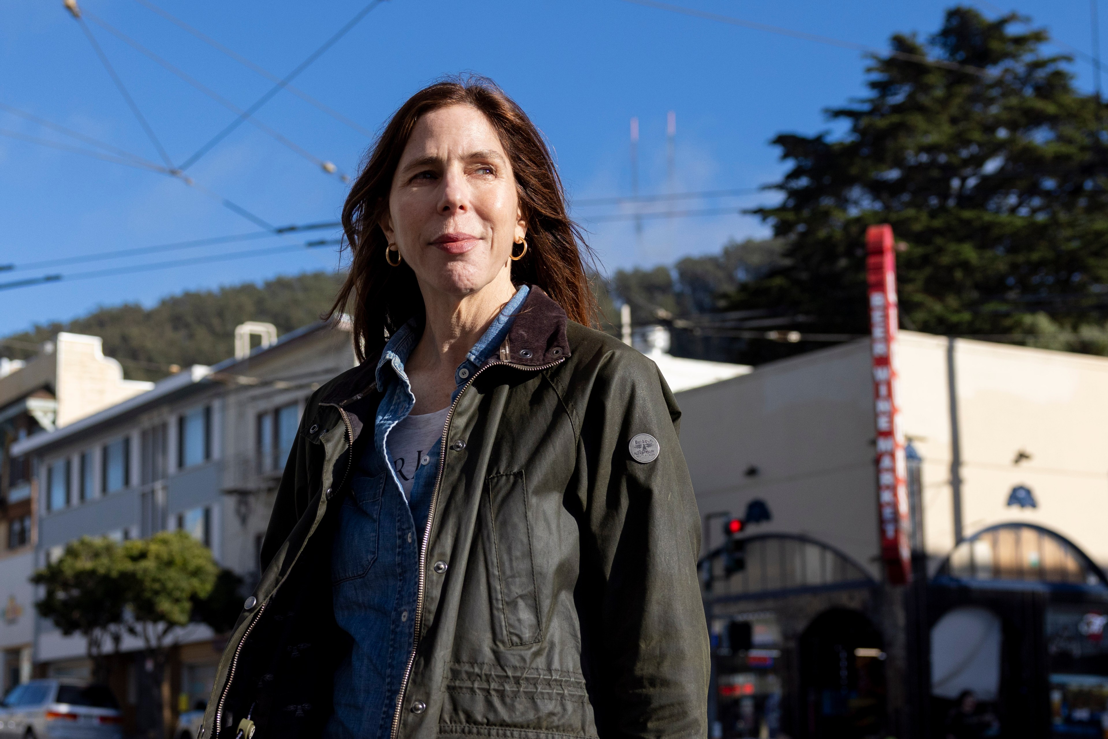 A woman in a green jacket stands outdoors with a backdrop of buildings, trees, and a clear blue sky. She gazes slightly upward and appears thoughtful.