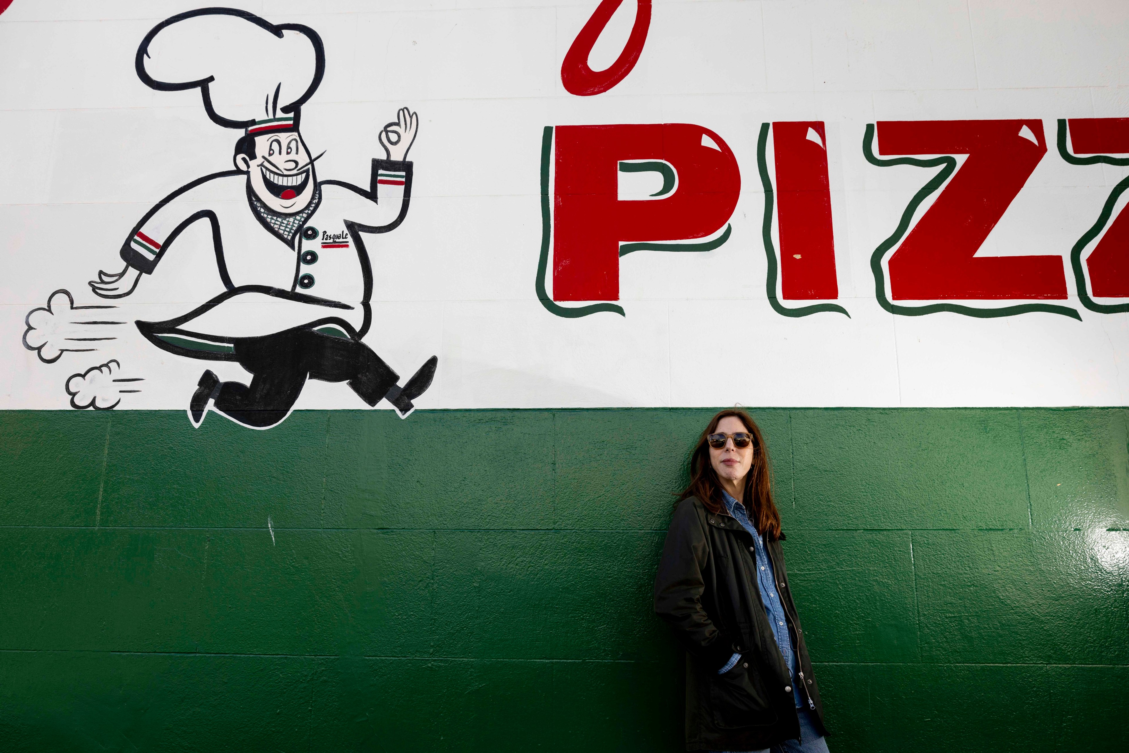 A woman in sunglasses leans against a green wall featuring a large mural of a smiling chef and the word &quot;PIZZA&quot; in bold red letters.