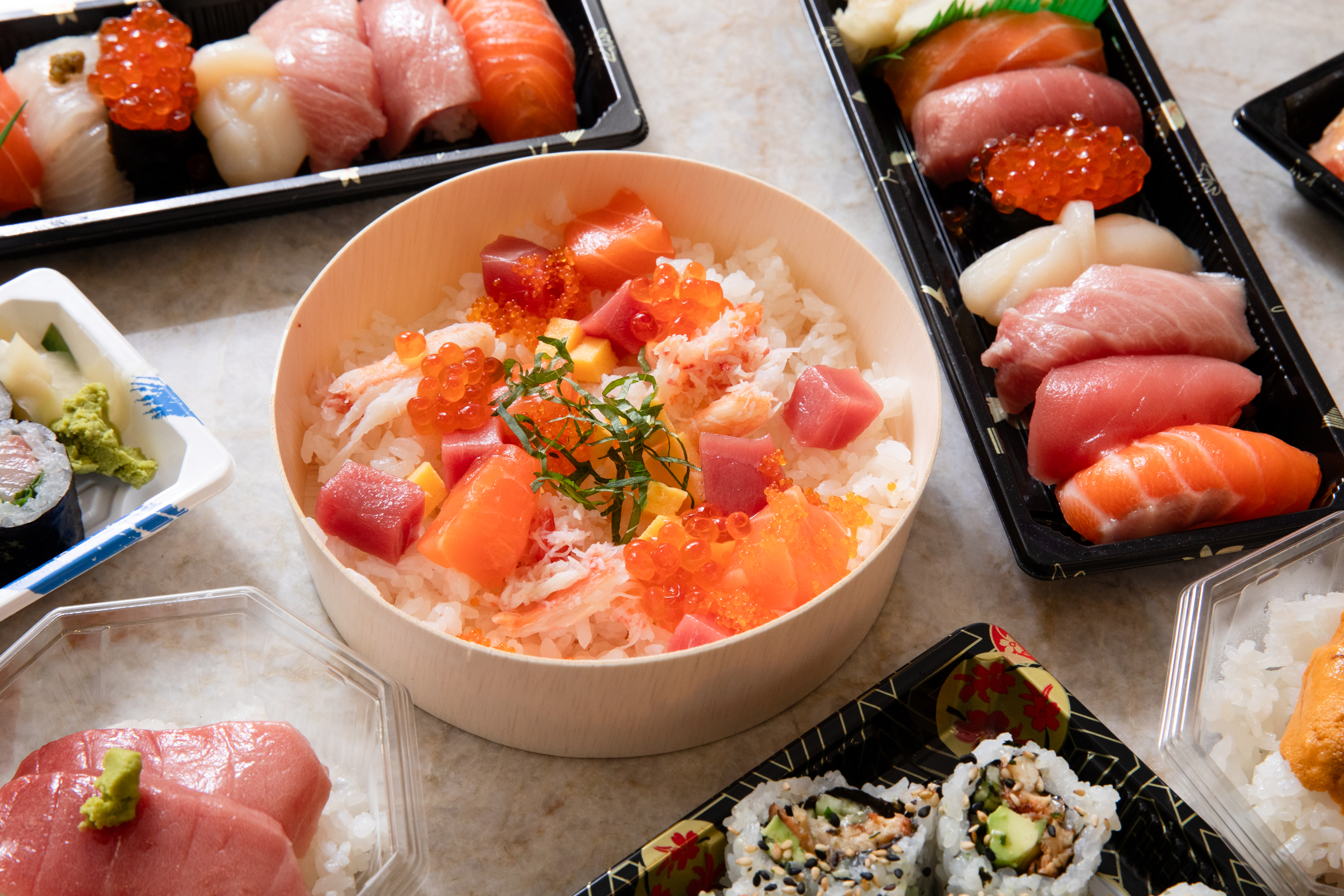 This image shows a variety of sushi dishes. At the center is a round bowl of rice topped with diced raw fish, roe, and seaweed. Surrounding it are trays of various sushi.