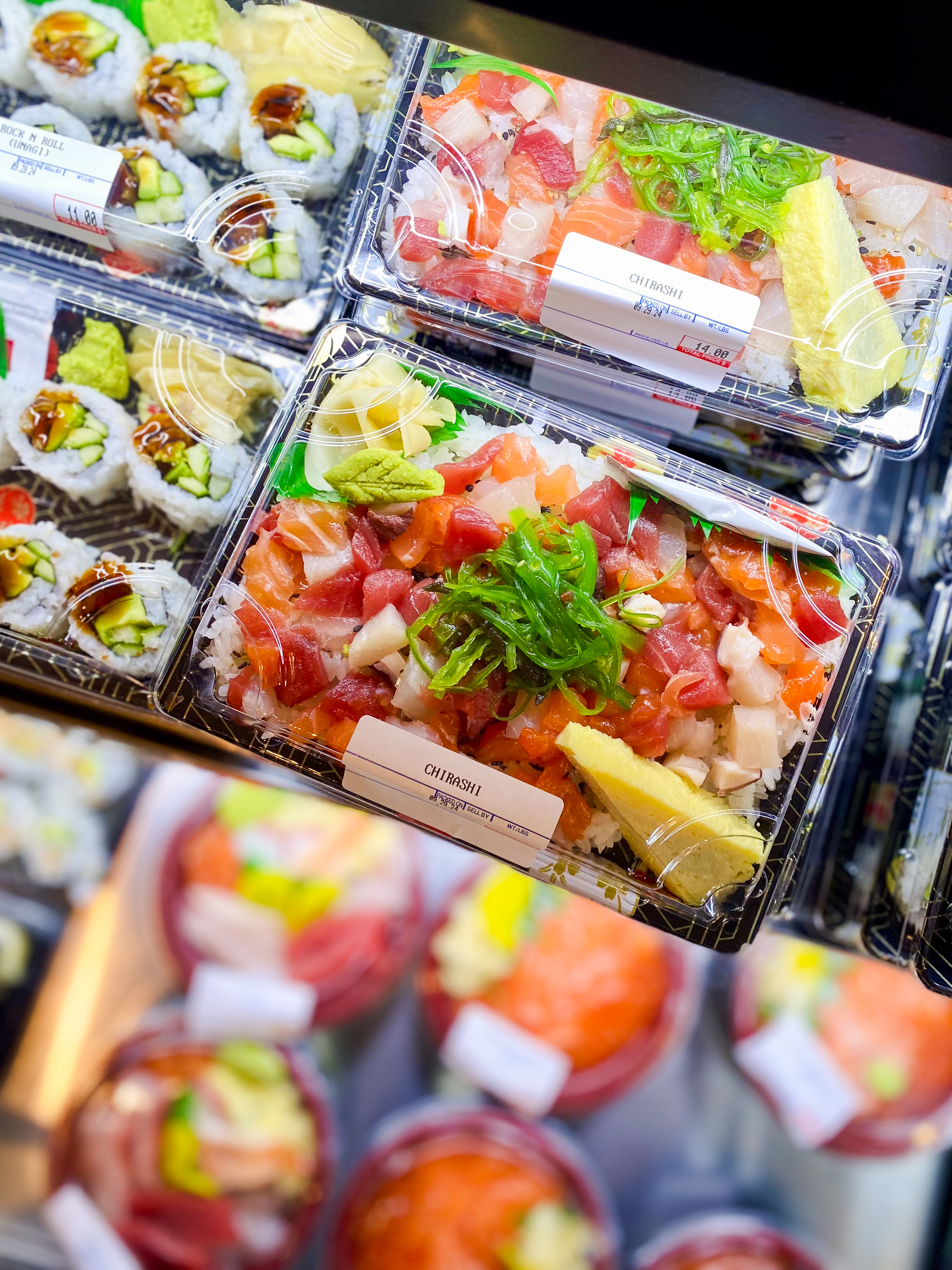 The image shows packaged sushi and sashimi boxes in a display case, with colorful assortments of fresh fish, rice, and garnishes, neatly arranged and labeled.