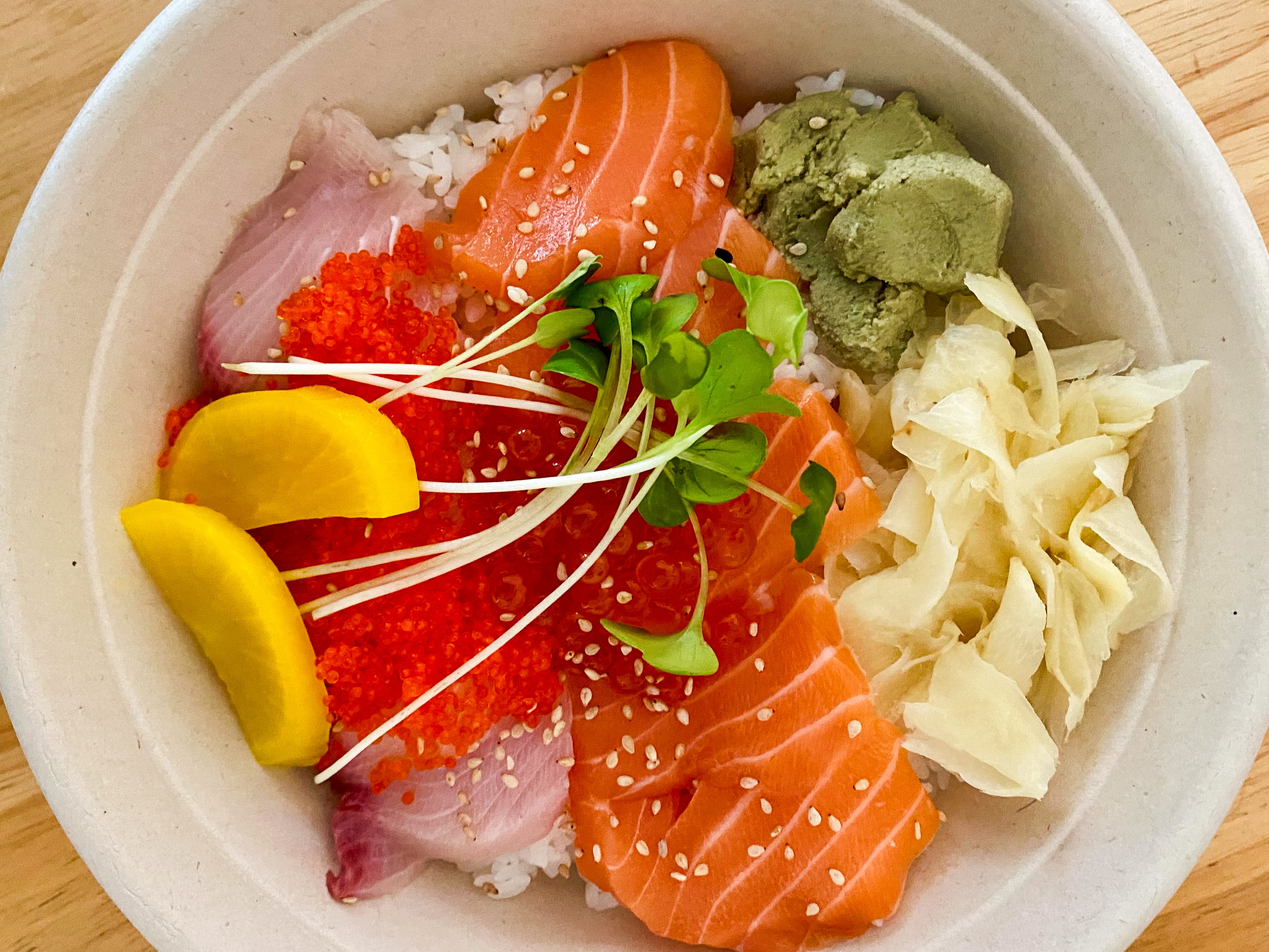 A bowl of sushi rice topped with sliced raw salmon, yellow pickled radish, fish roe, wasabi, ginger, and sprouts.