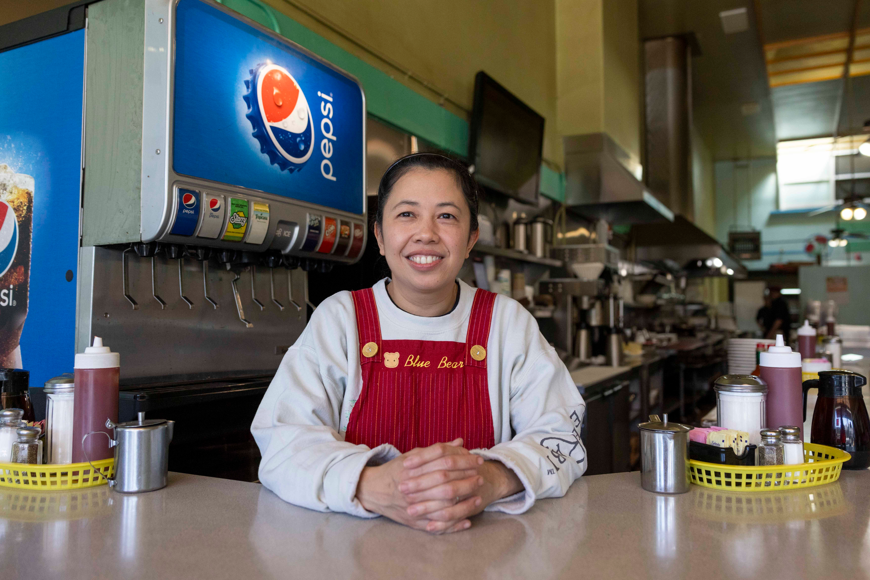 A smiling woman in red overalls and a white shirt stands behind a counter in a diner, with a Pepsi soda dispenser and various condiments around her.