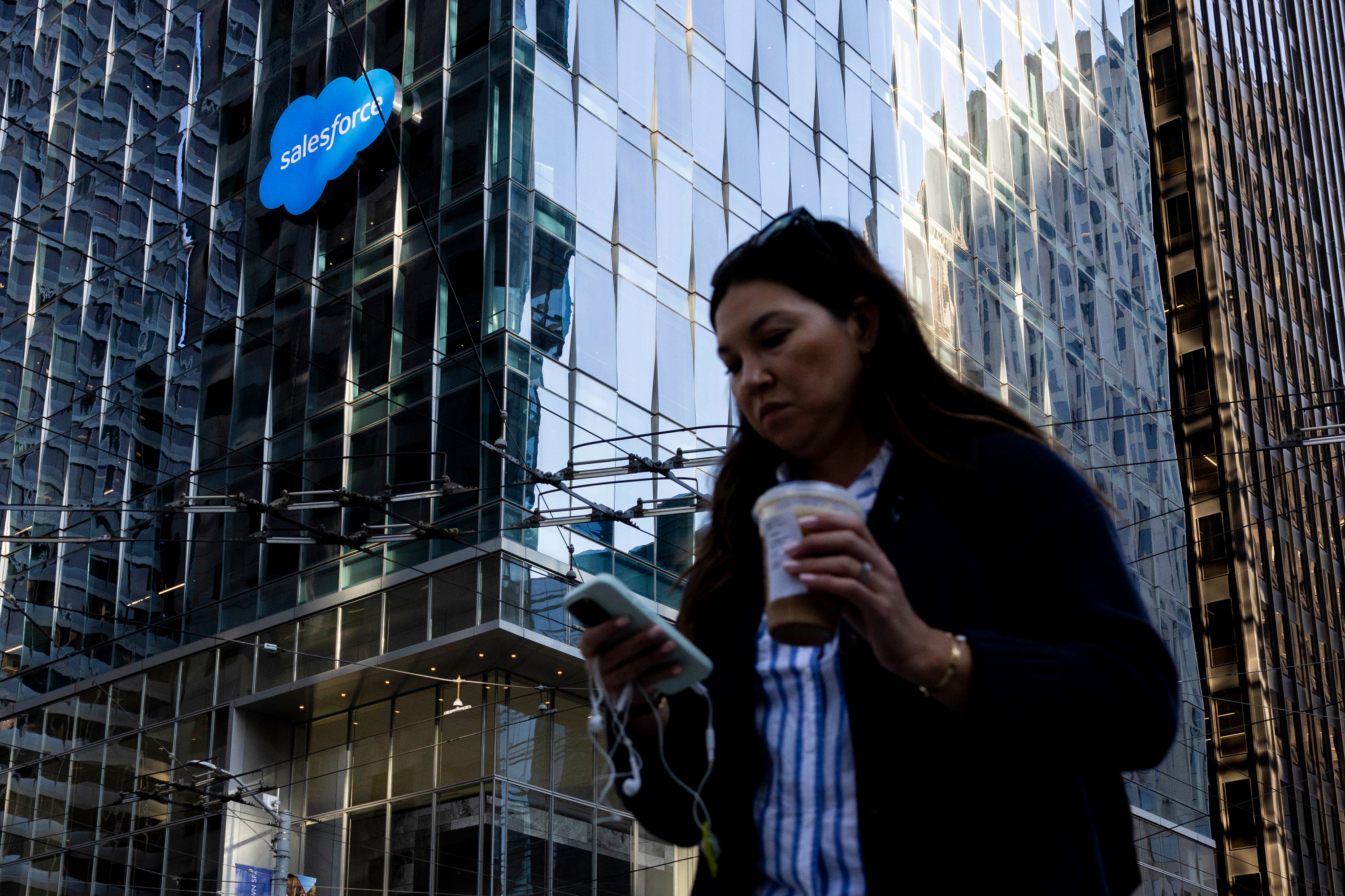 A woman is walking while looking at her phone and holding a drink. Behind her is a glass building with a Salesforce logo.