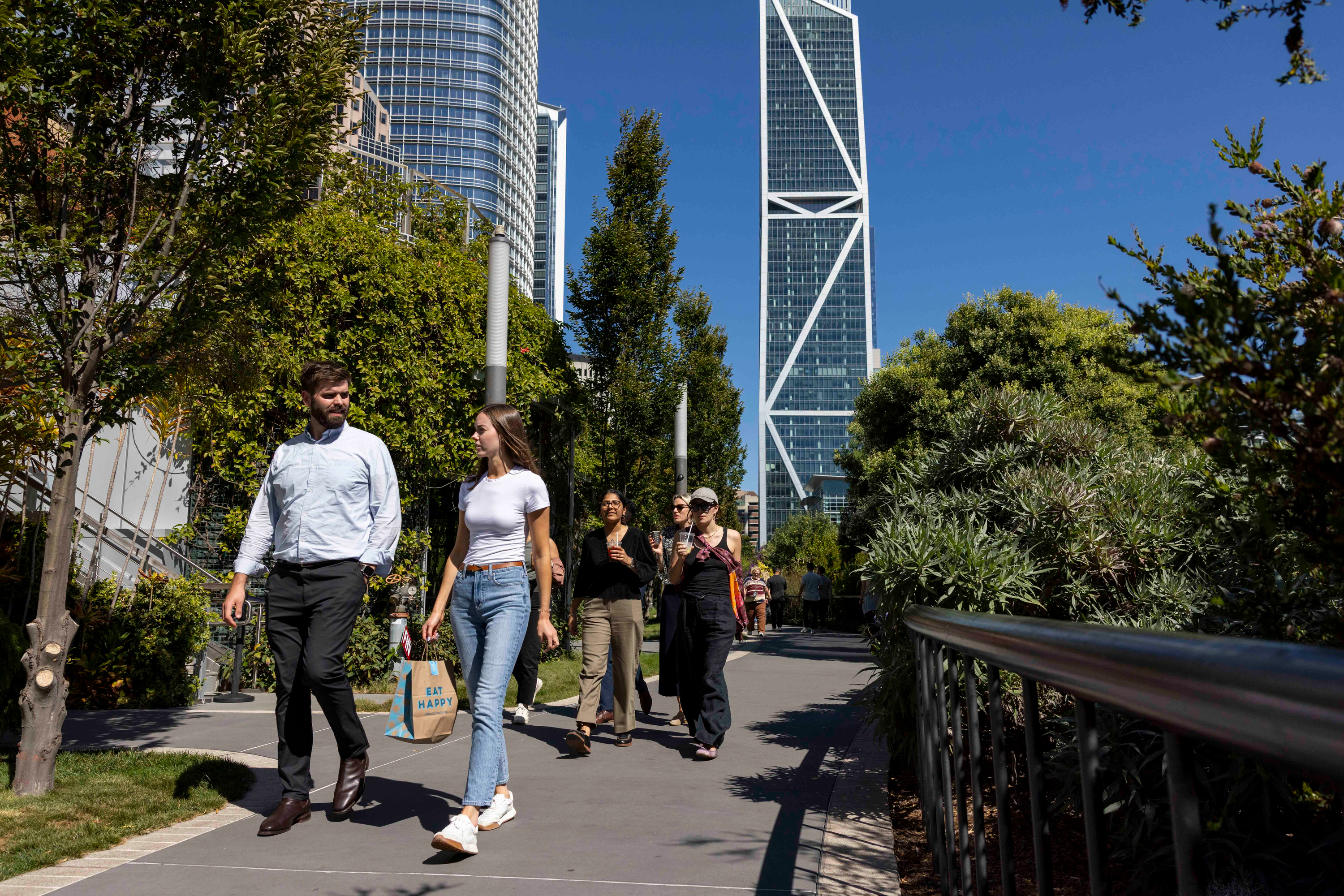 A diverse group of people are walking on a sunny, tree-lined path with a tall, modern skyscraper in the background, under a clear blue sky.