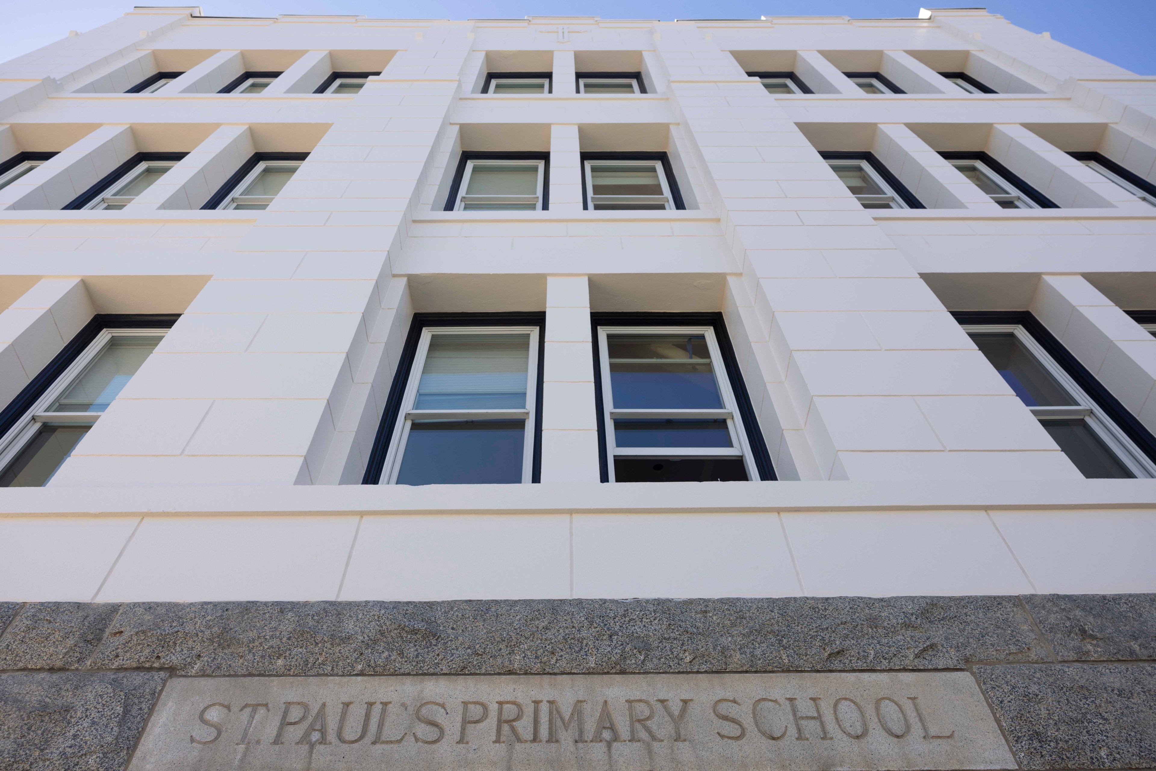 The image shows a white building with a modern design featuring large windows. The stone plaque at the base reads &quot;St. Paul's Primary School.&quot;