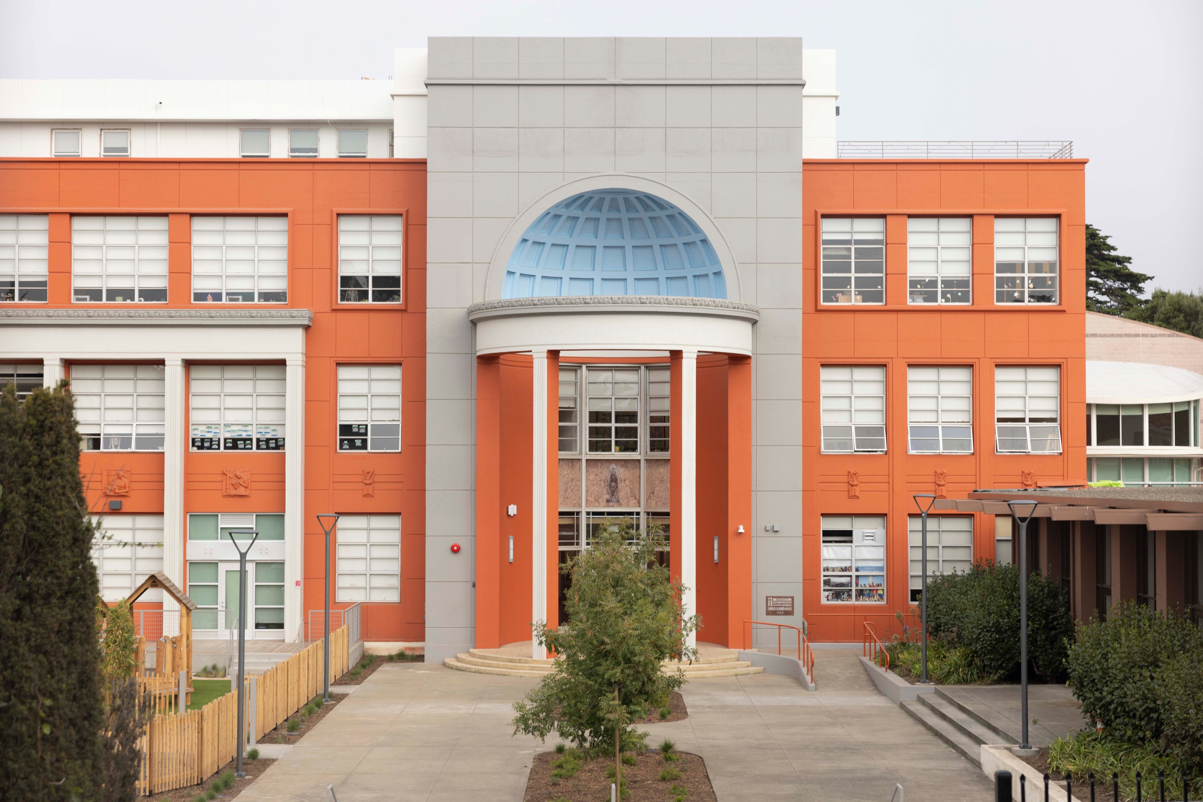 The image shows a modern building with an orange and gray facade, featuring large windows and a prominent arched entrance, surrounded by greenery.