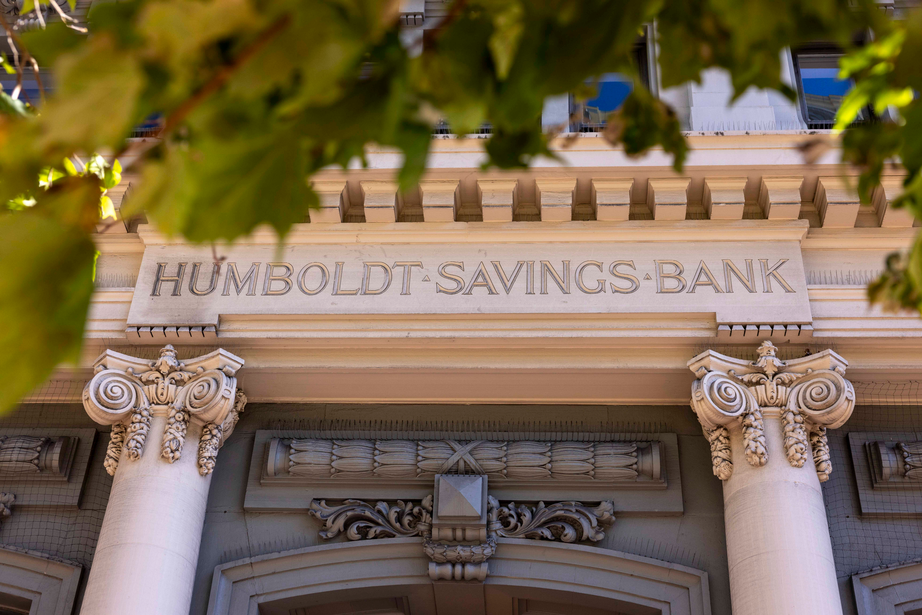 The image shows the facade of Humboldt Savings Bank, featuring classical architectural elements like ornate columns and intricate stone detailing, partially framed by tree leaves.