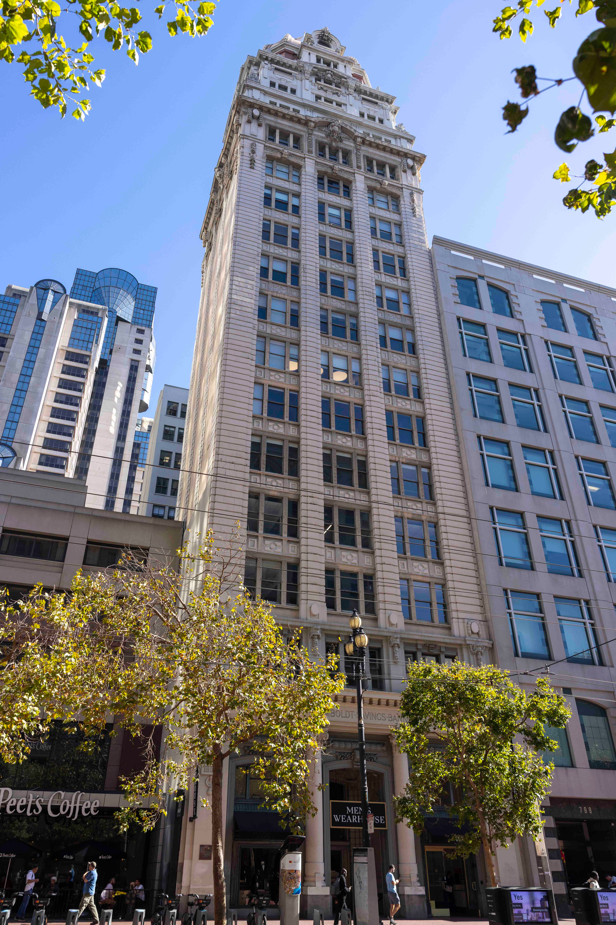 A tall, ornate building with numerous windows and decorative details stands among other high-rises under a clear blue sky, with trees and pedestrians on the street below.