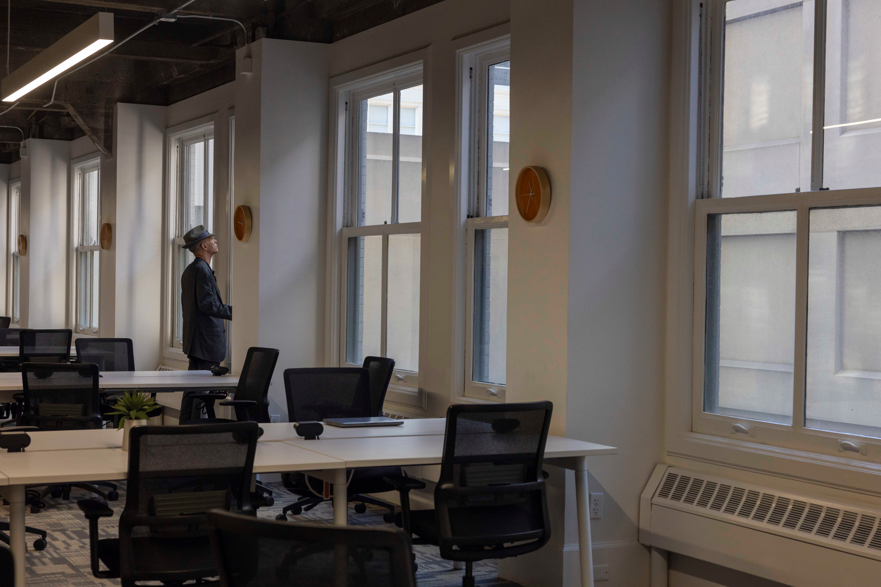 A man in a suit and hat gazes out of large windows in a modern, empty office with rows of desks and chairs. The space is bright with natural light.