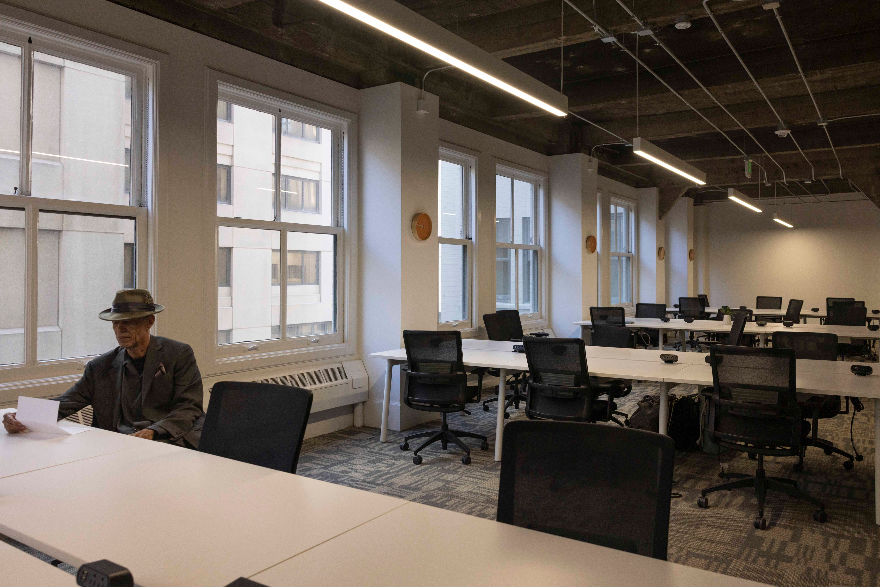 A man in a hat sits reading at a desk in an empty office with several rows of desks and chairs, large windows, and overhead lights.