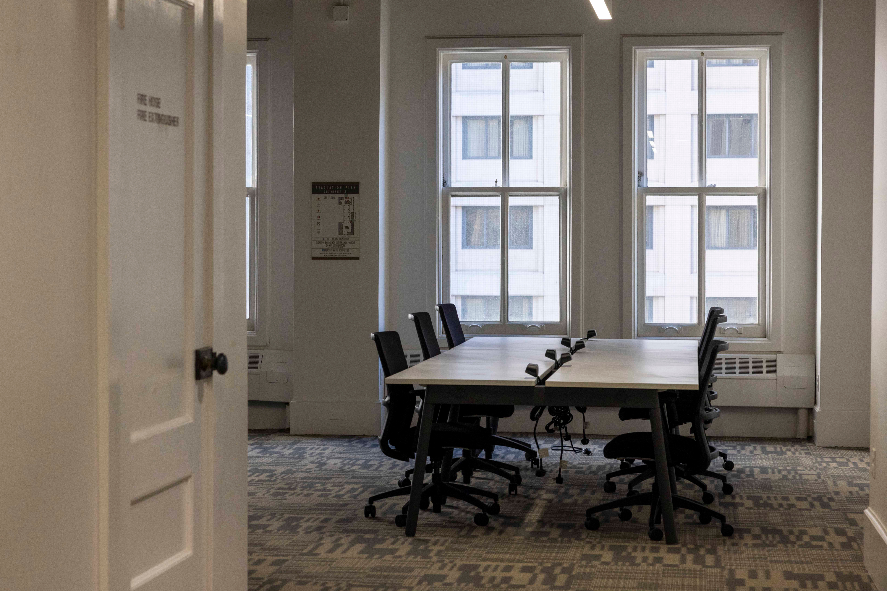 The image shows a clean, modern conference room with a long table and eight black office chairs, facing three large windows. The walls are white, and the room is well-lit.