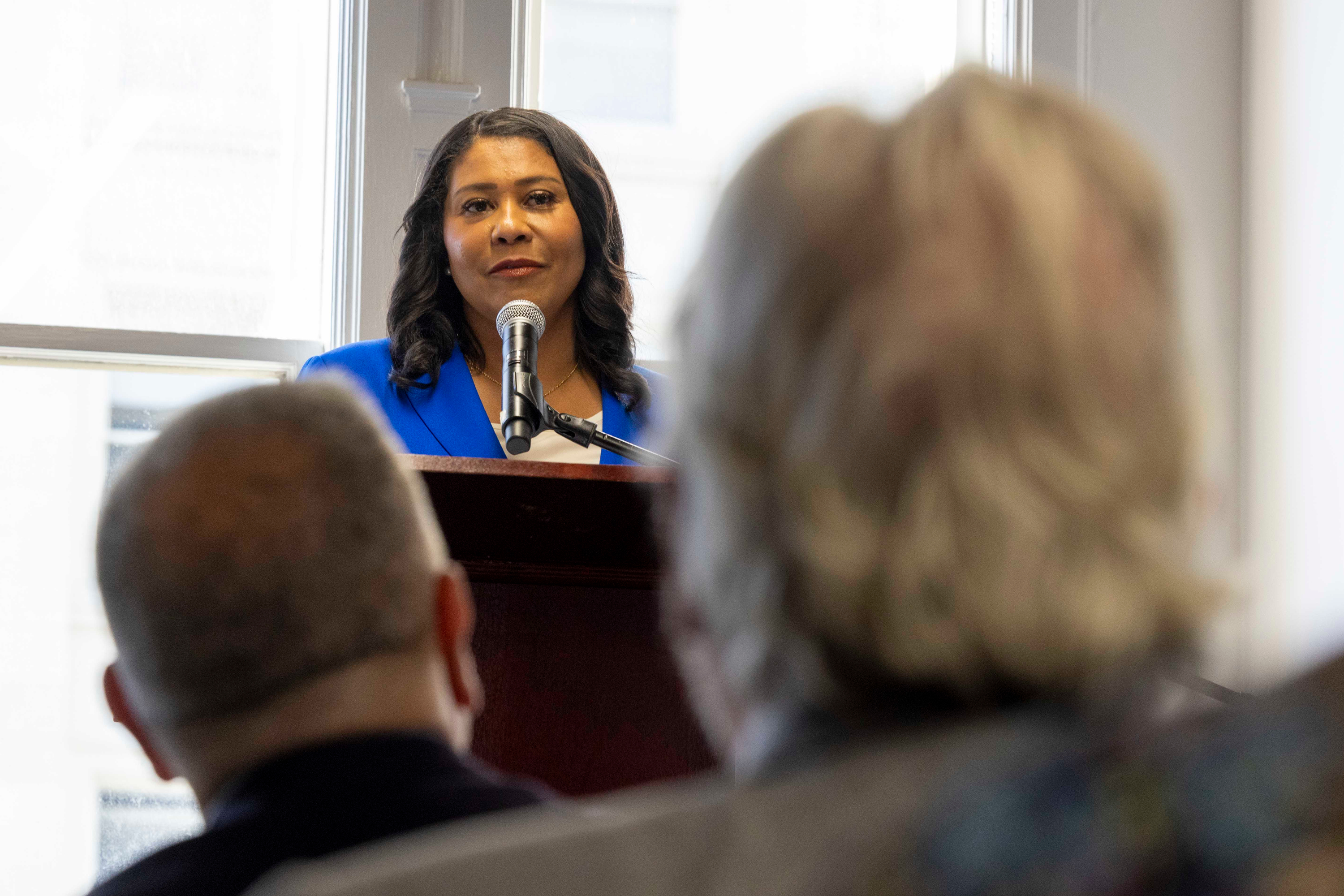 A woman in a blue blazer speaks at a podium in front of a microphone, while an audience listens attentively. The setting appears to be indoors.