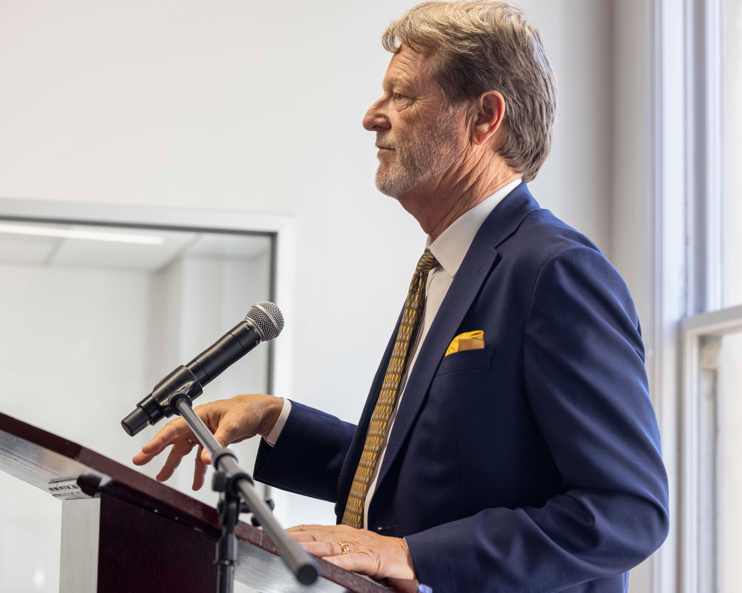 A man in a navy suit and yellow tie speaks at a podium with a microphone. He has gray hair and a beard, and the background shows a white room with a window.