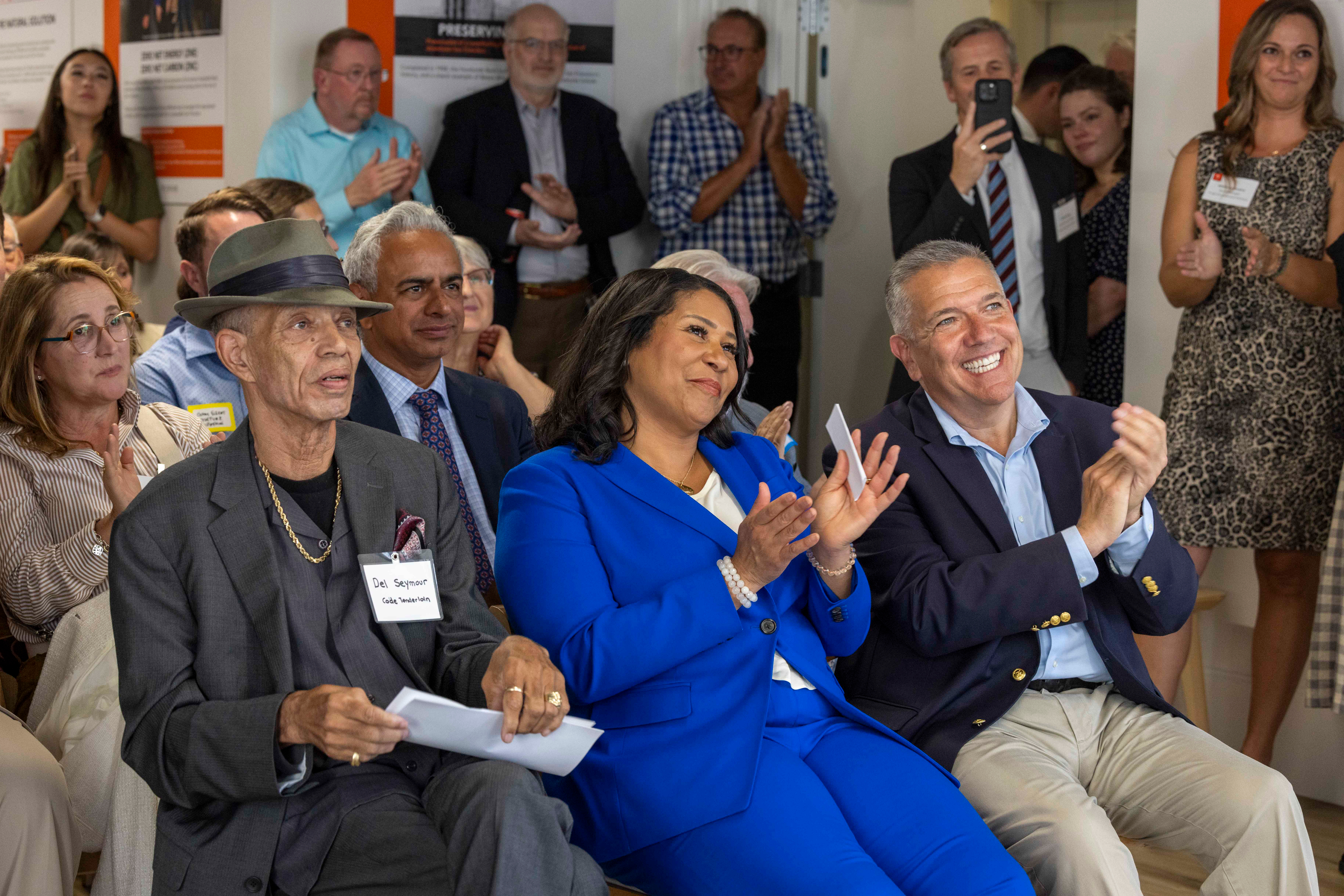 A diverse group of people, including a man in a hat and a woman in a blue suit, sit and stand, smiling and clapping in a well-lit room.