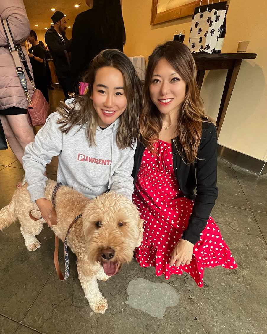 Two women are smiling; one wears a &quot;Pawrent&quot; hoodie, and the other a red polka-dot dress. They're squatting next to a fluffy, happy dog indoors.