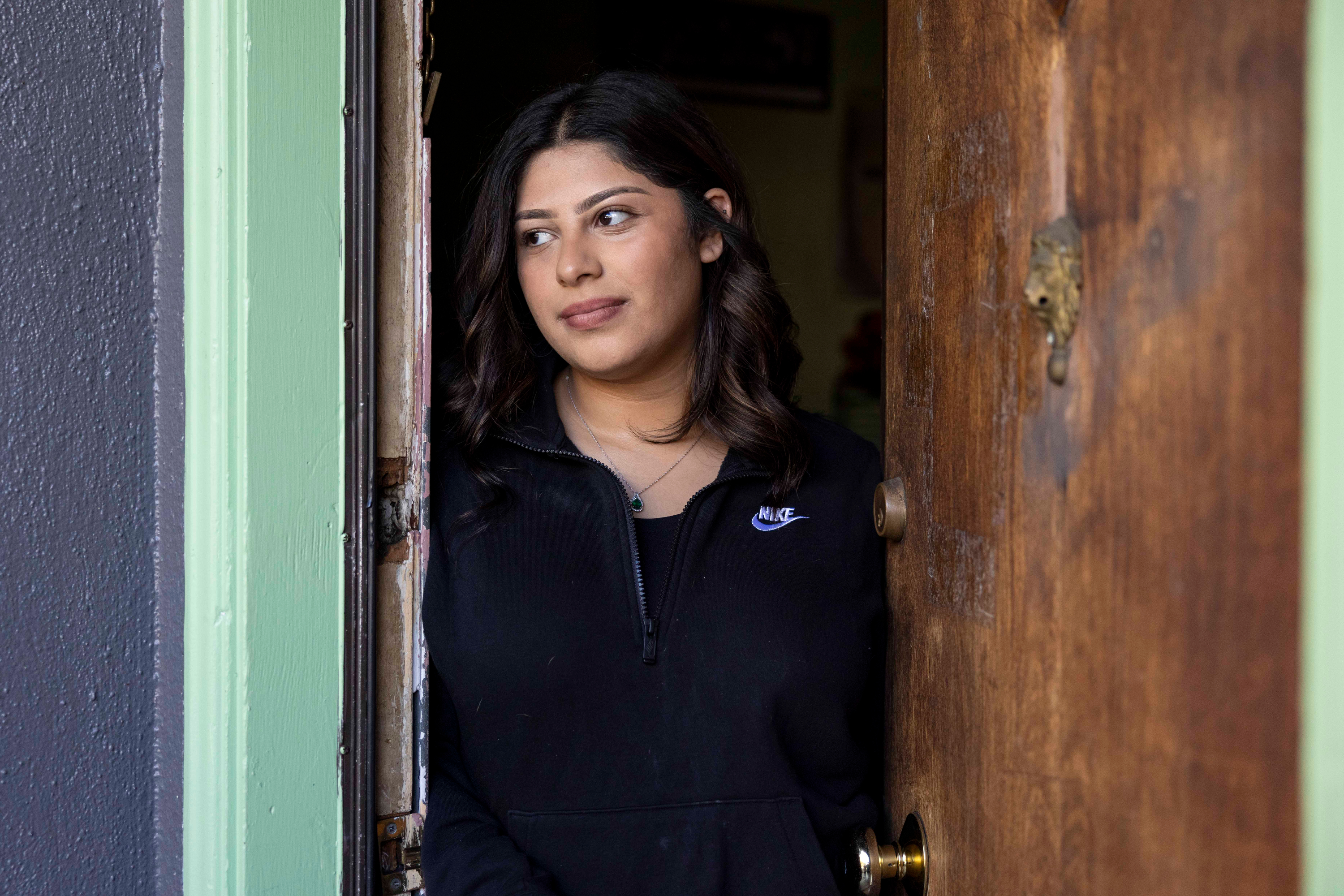 A woman with wavy brown hair, wearing a black zip-up sweater, stands in a doorway, partially behind a wooden door, looking to her right with a calm expression.