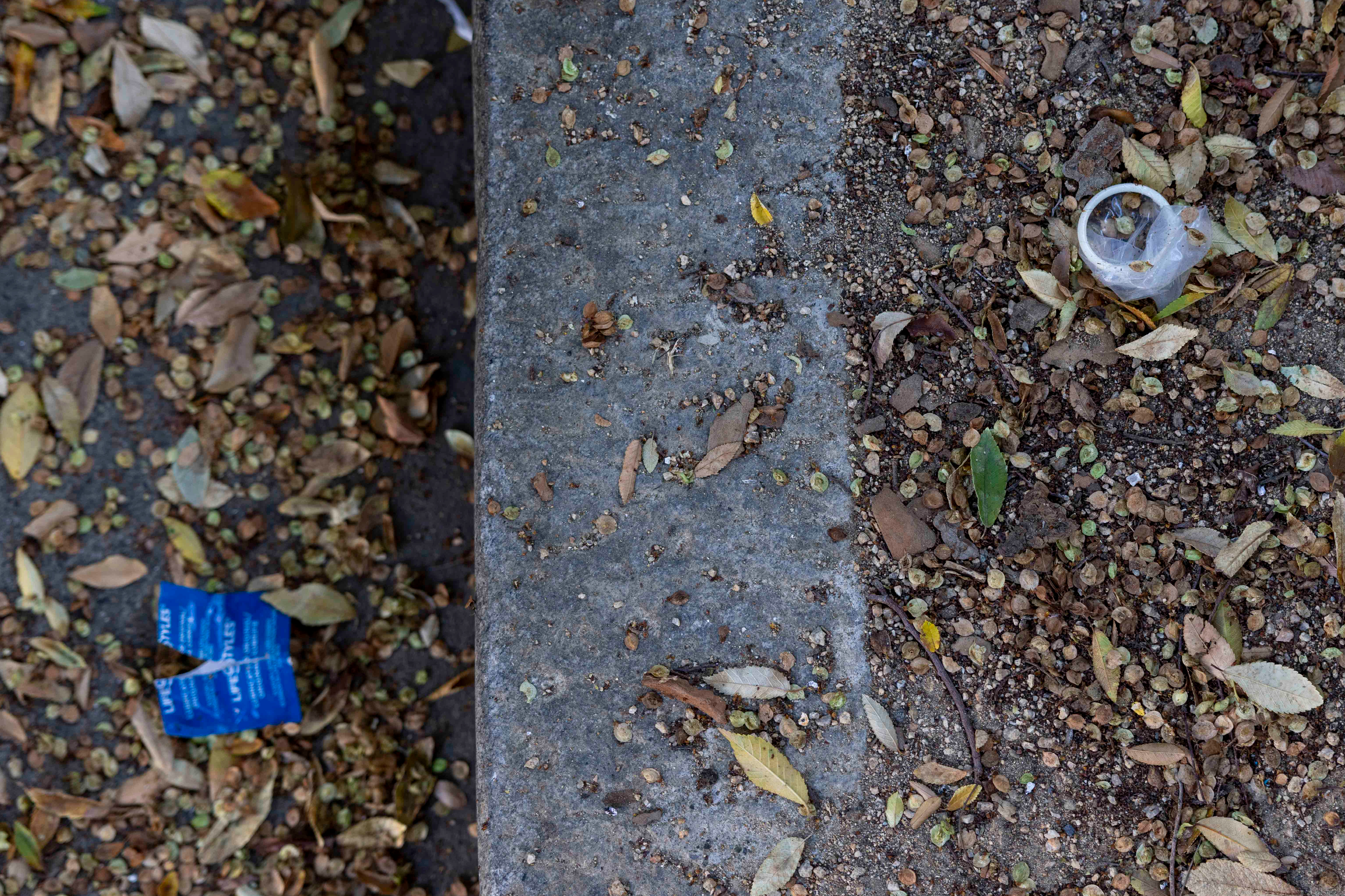 The image shows a ground scattered with dried leaves and debris. A torn blue condom wrapper and a used condom are visible among them.