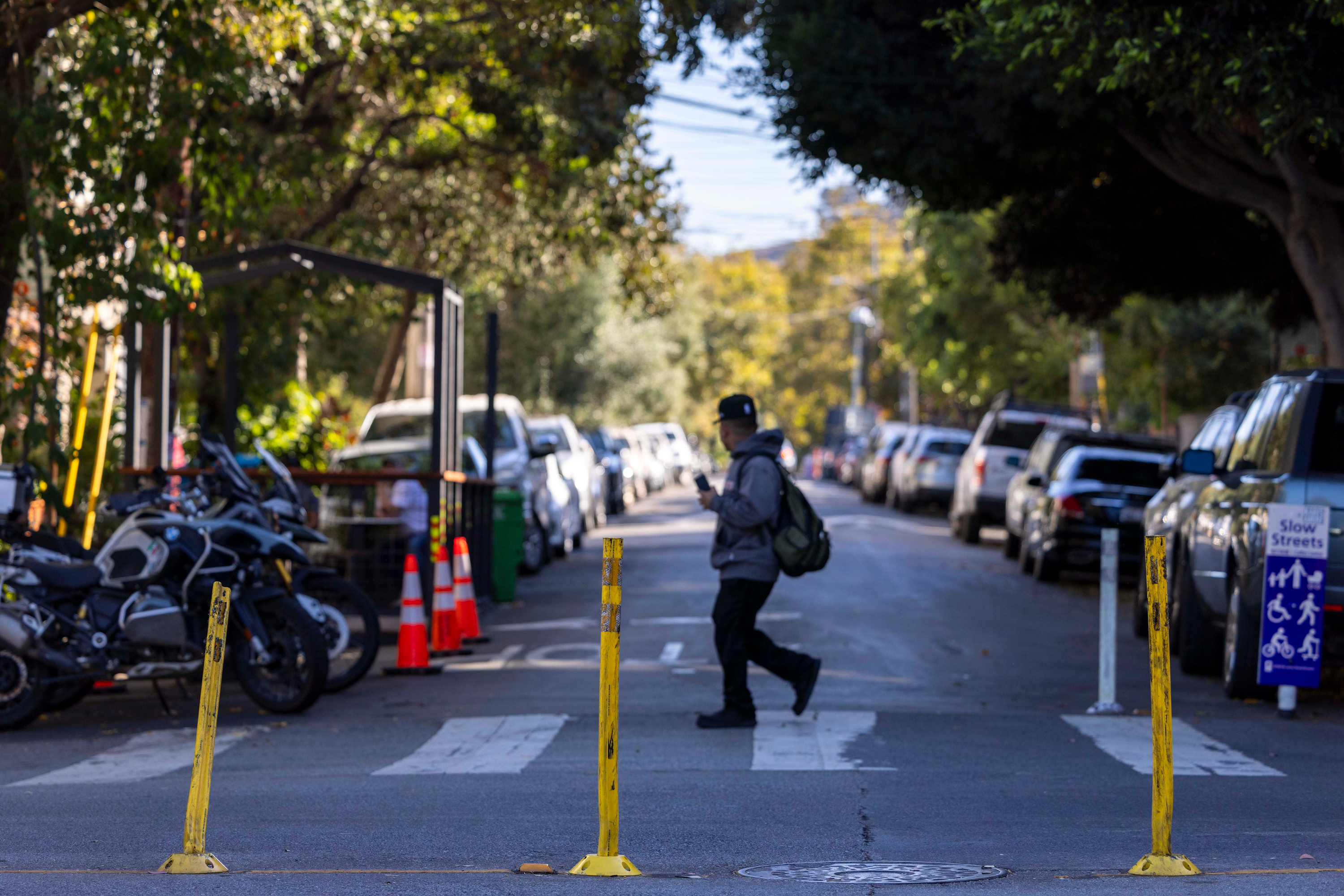 A person crosses a tree-lined street with parked cars and motorcycles. Yellow poles and road cones are present, and a &quot;Slow Streets&quot; sign is visible on the right.